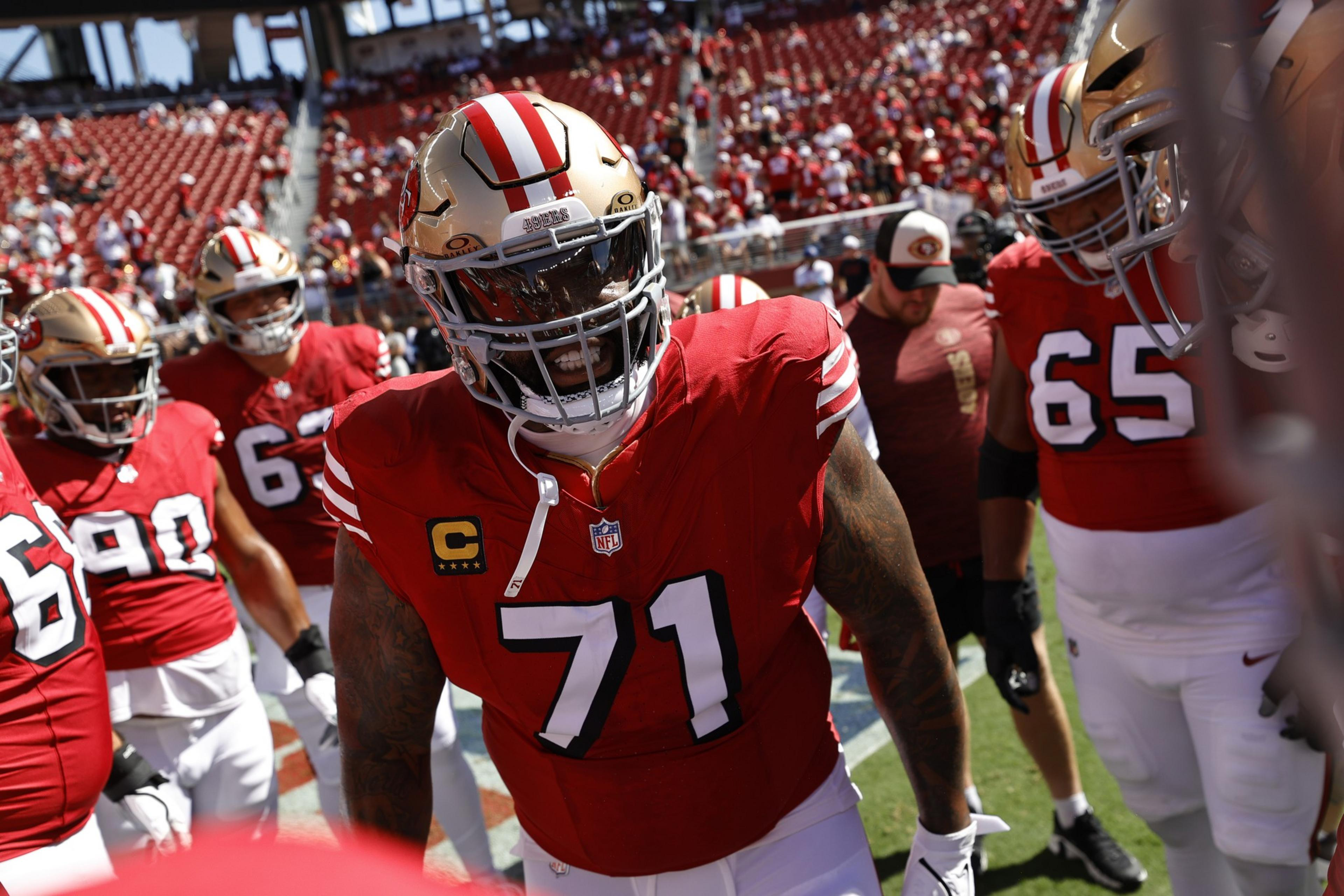 A football player wearing a red jersey with number 71 and gold helmet stands among teammates in a stadium filled with fans.