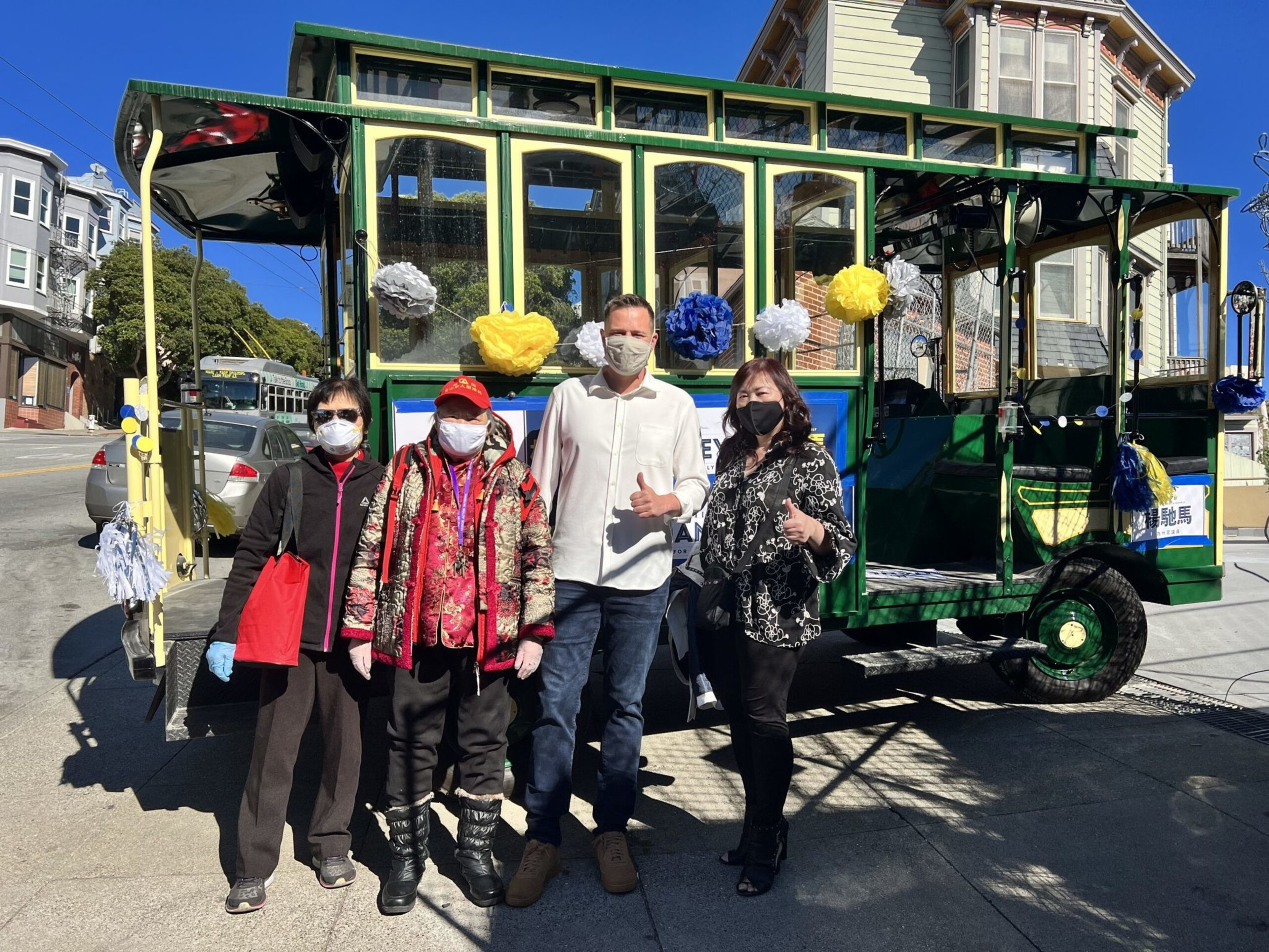 Four people wearing face masks stand in front of a green and yellow decorated trolley on a sunny street.
