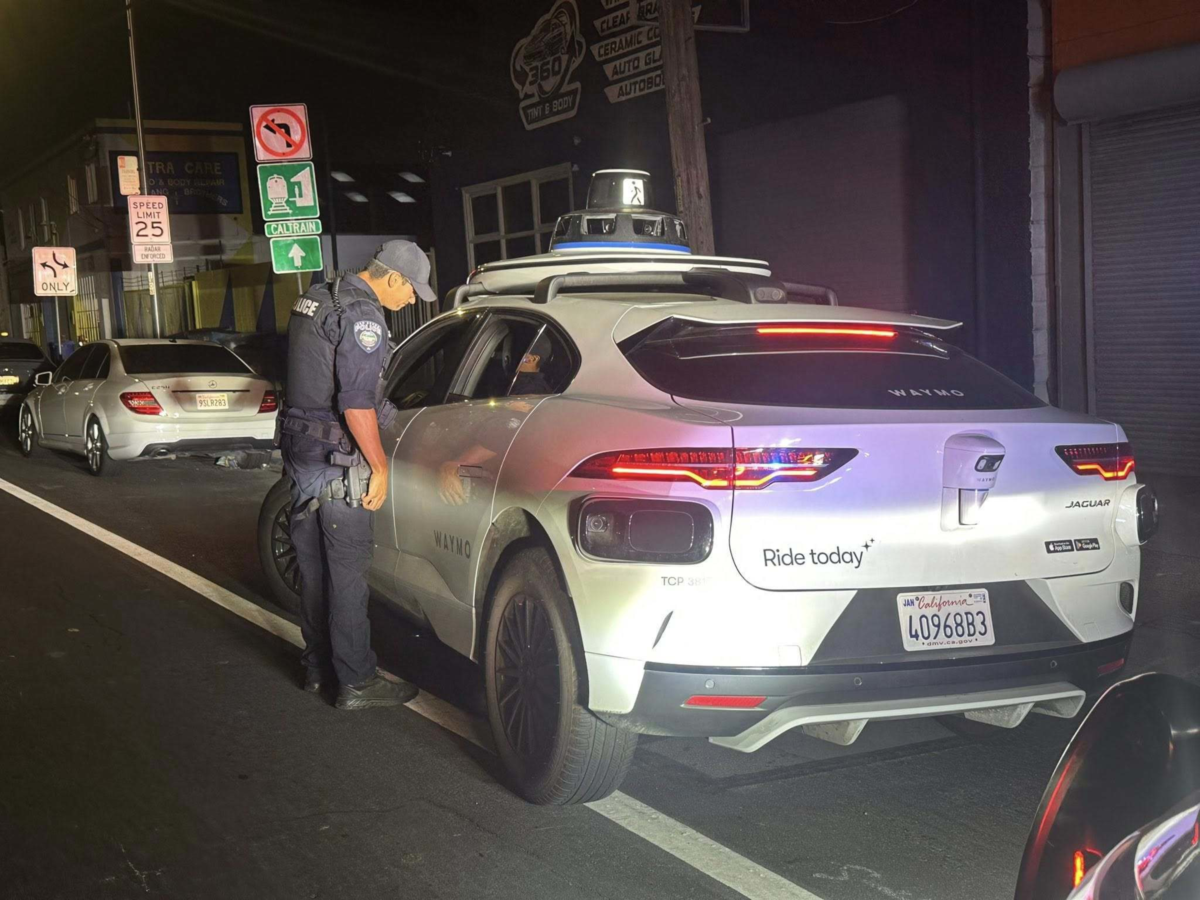 A police officer is speaking to the driver of a white Waymo Jaguar parked on the street at night.