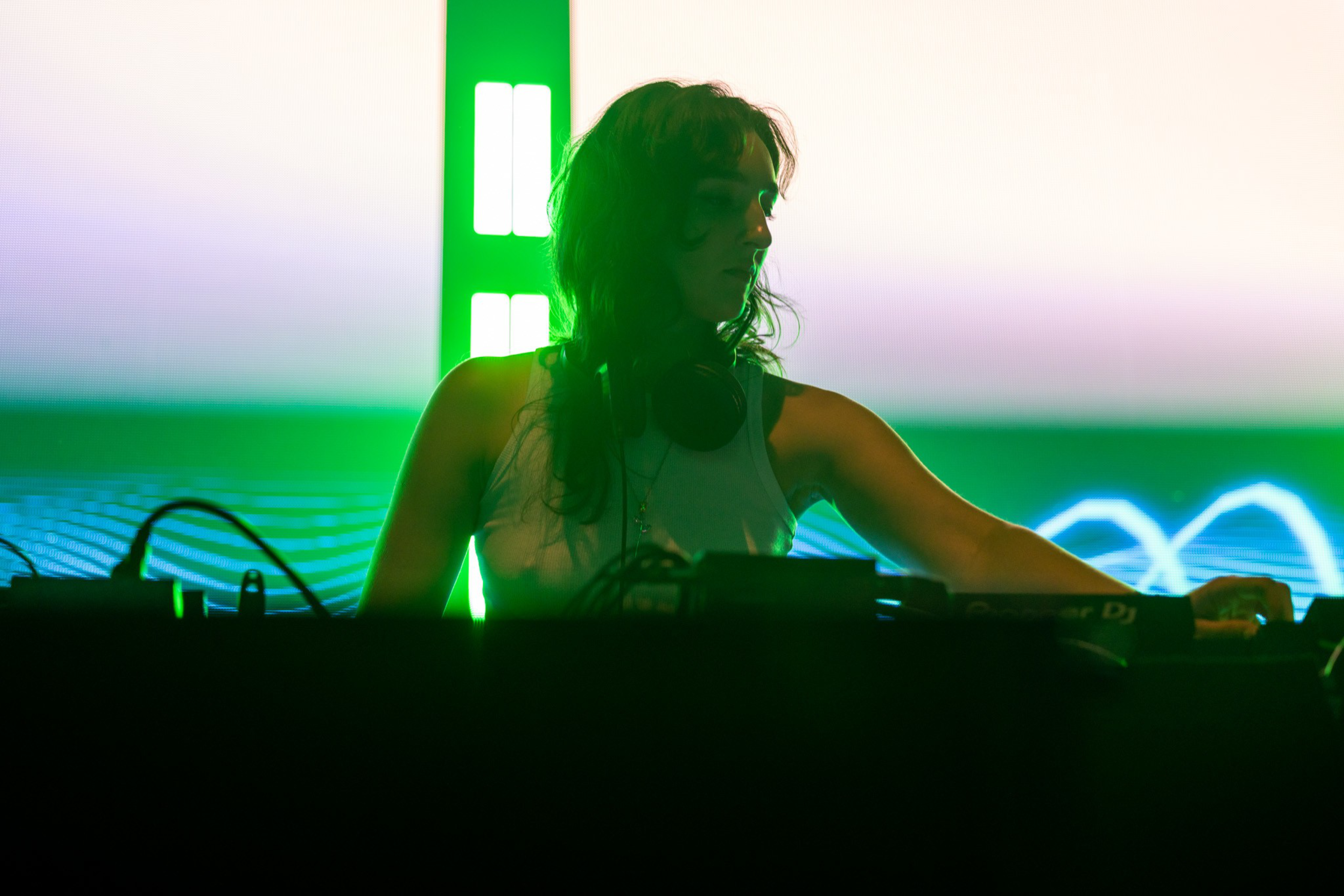 A DJ wearing headphones and a tank top is performing against a glowing green and white background with electronic equipment in front.