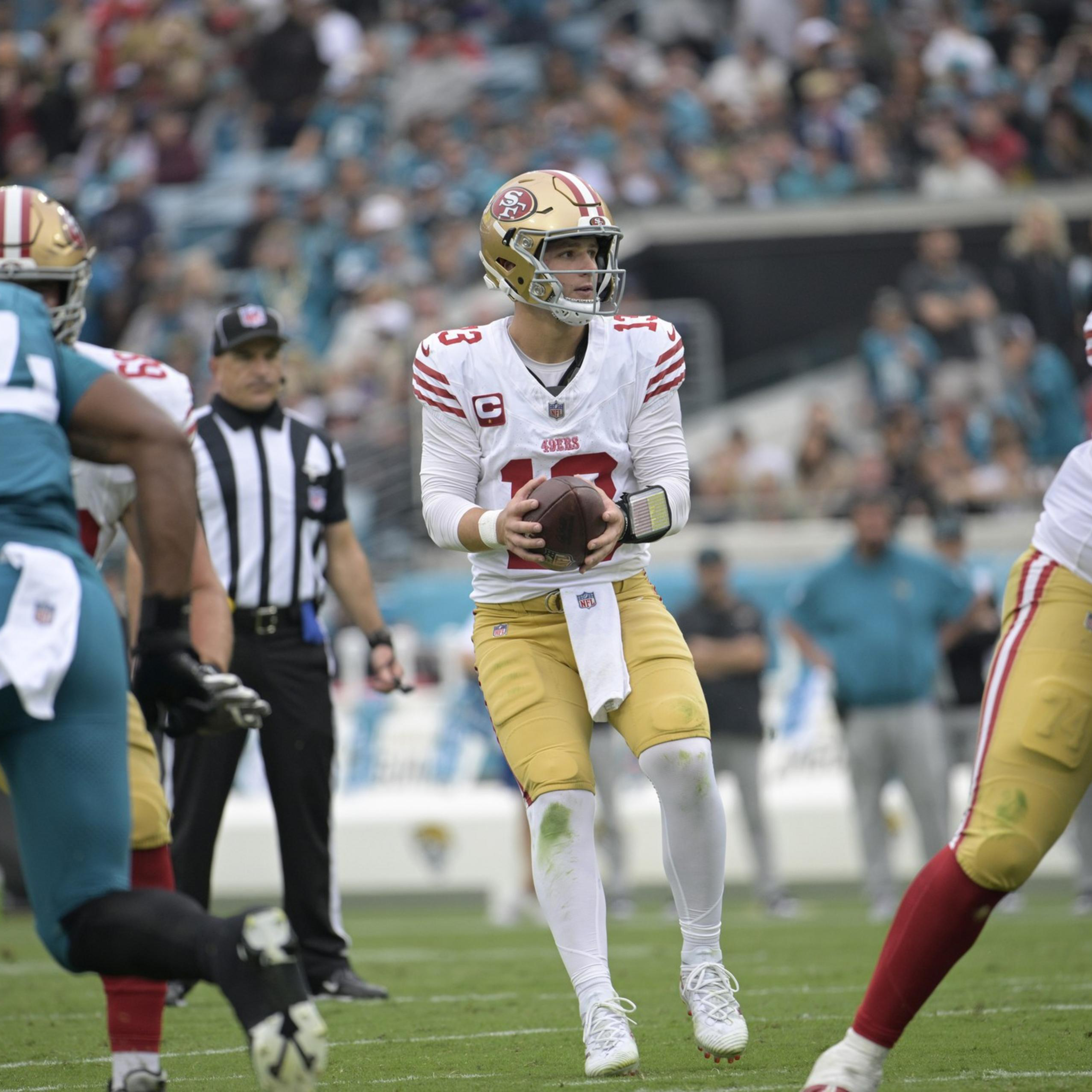 A football quarterback in a white and red uniform prepares to throw the ball while teammates block opponents wearing teal jerseys on a grassy field.