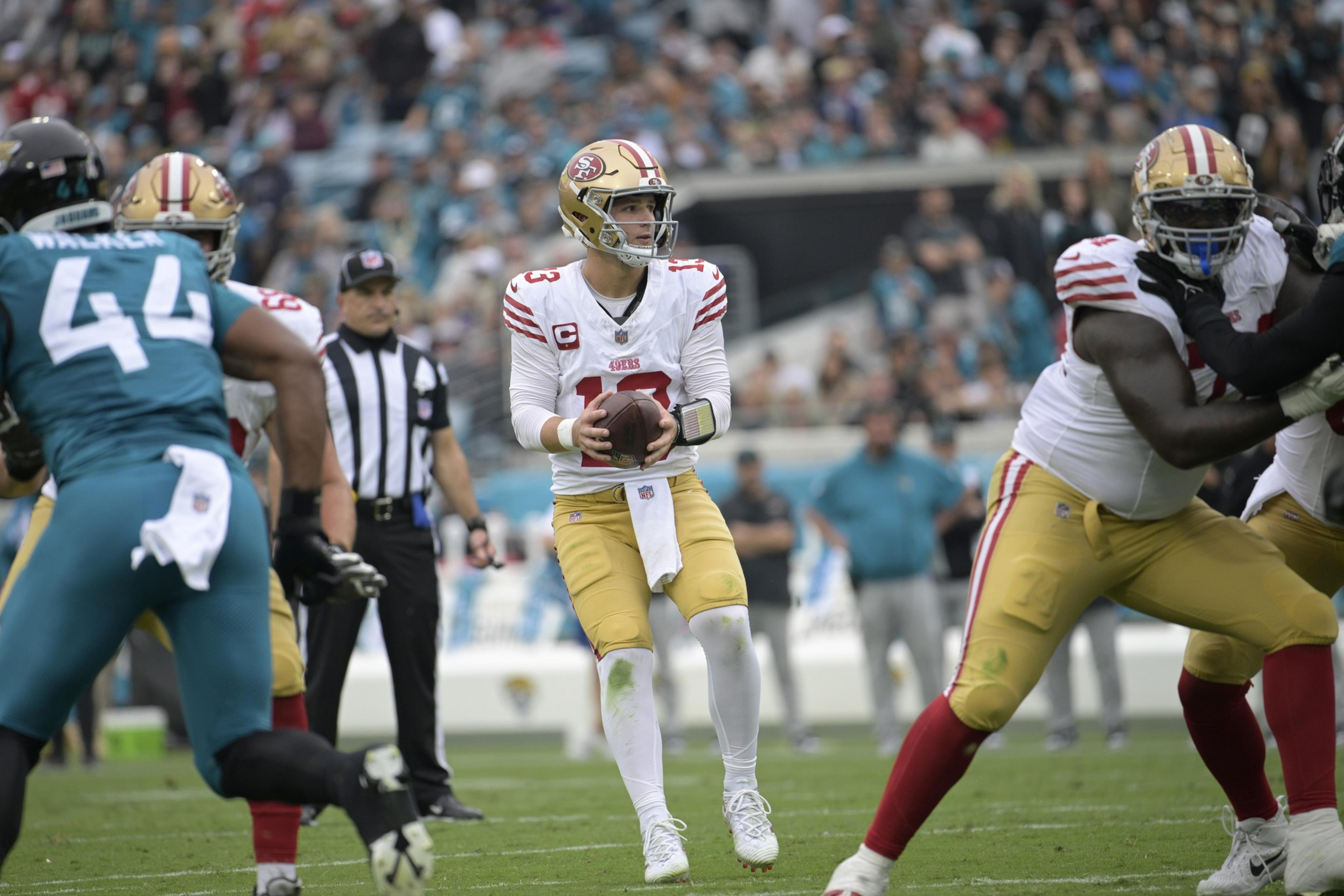 A football quarterback in a white and red uniform prepares to throw the ball while teammates block opponents wearing teal jerseys on a grassy field.