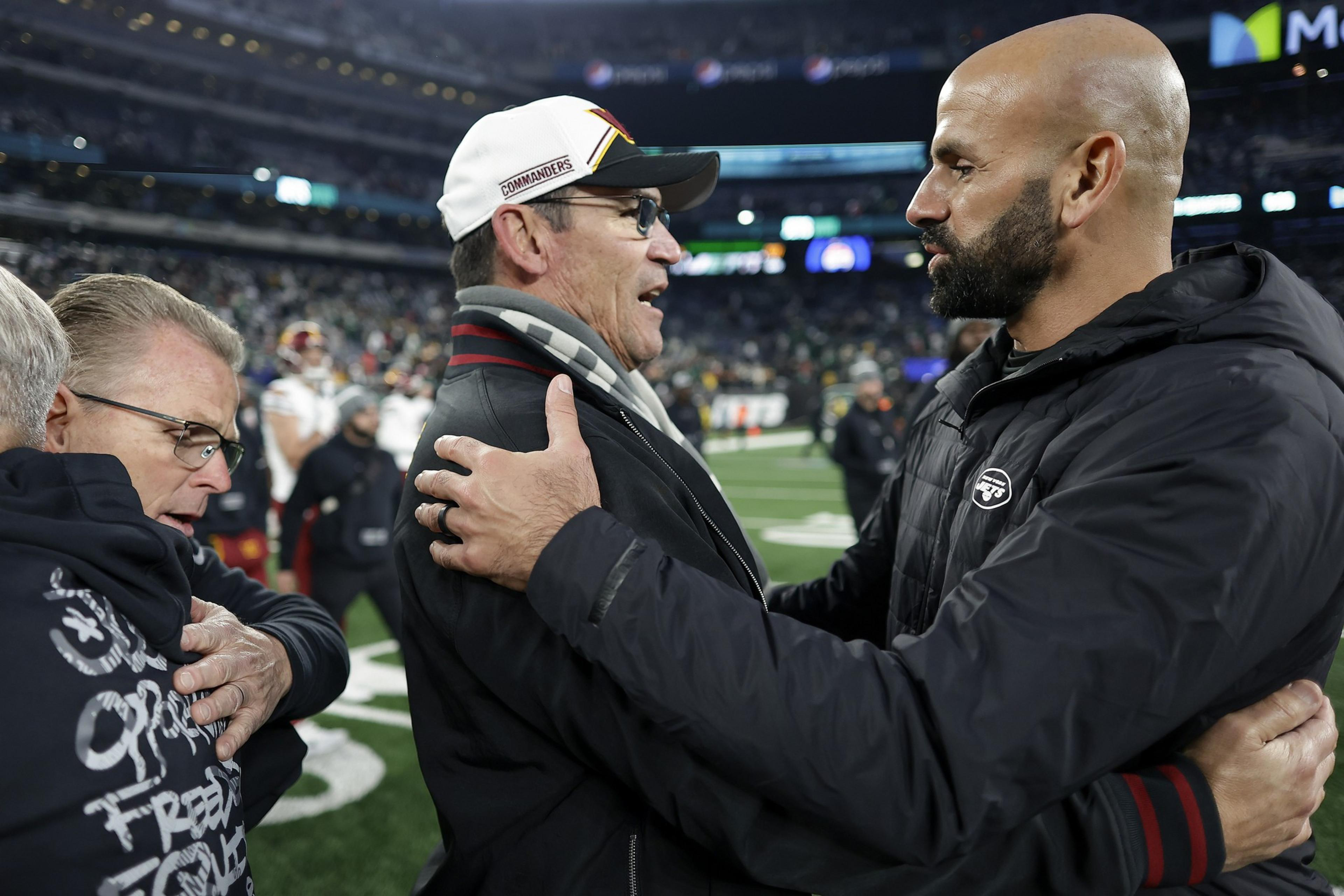 Two men warmly embrace on a football field, one wearing a “COMMANDERS” cap and glasses, the other bald with a beard in a black jacket.