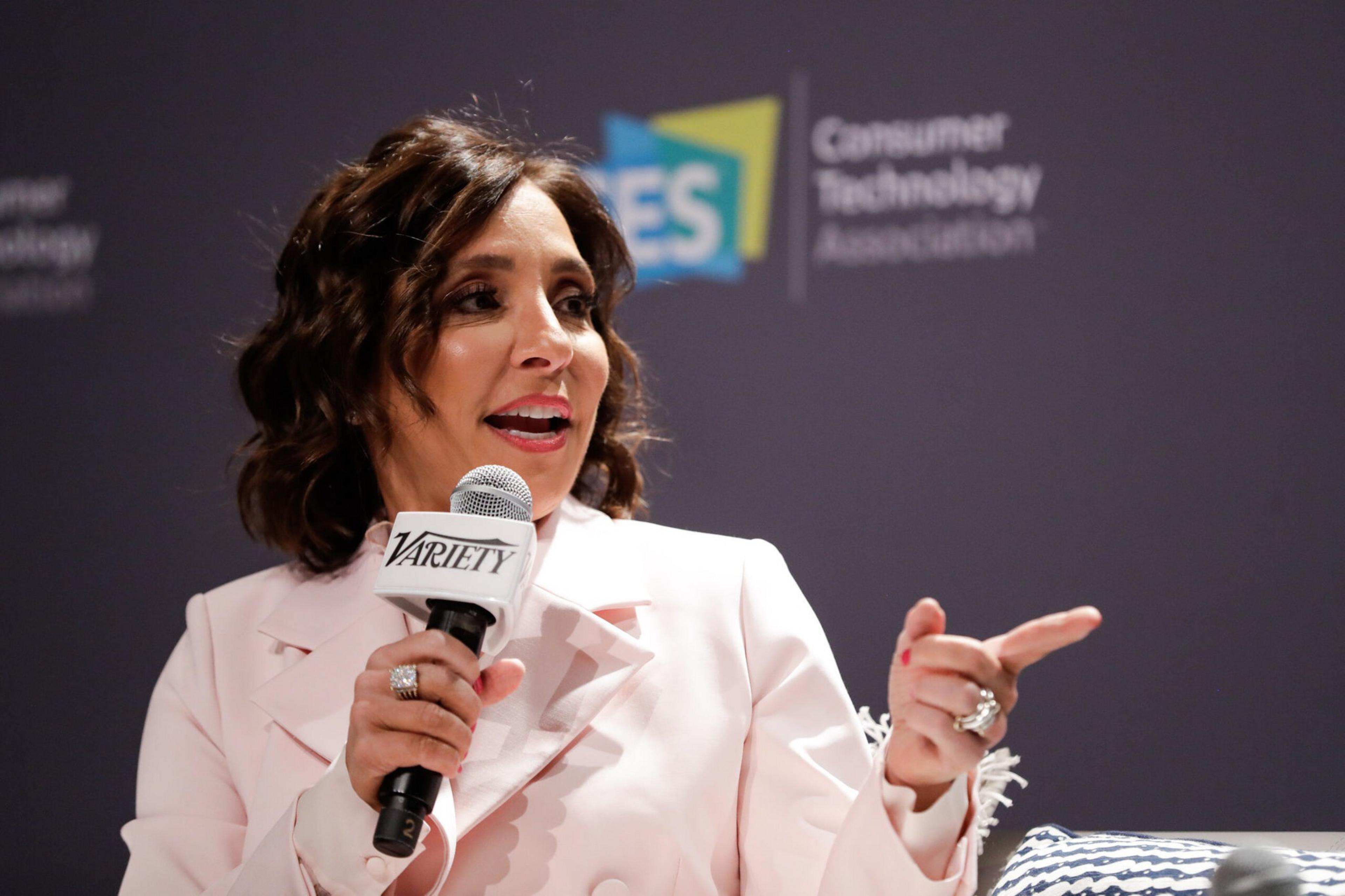A woman in a light pink suit holds a microphone labeled "Variety" and gestures with her right hand while speaking on stage.