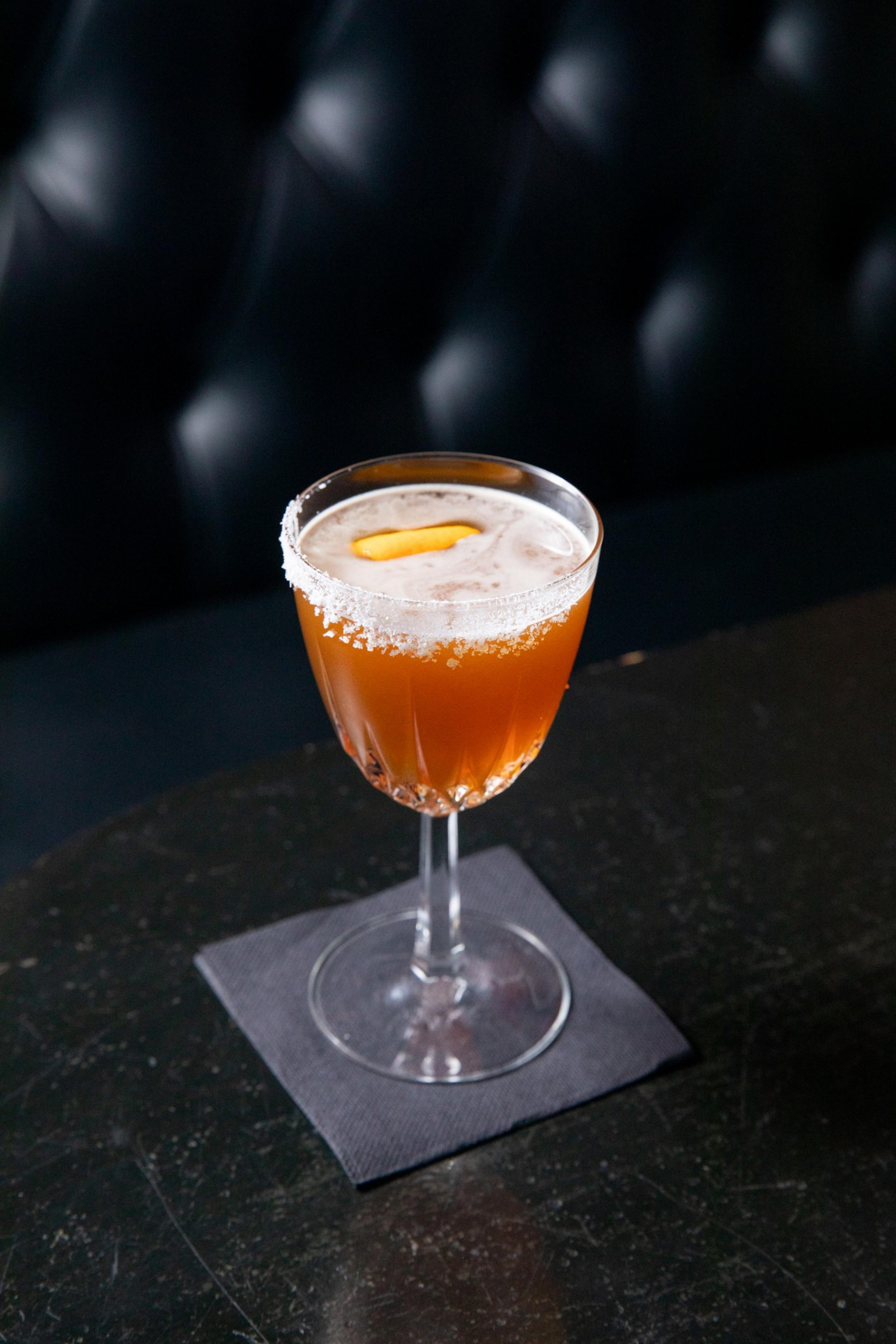 A cocktail in a fancy glass with a salted rim sits on a black napkin. The drink is amber-colored with a thin layer of foam on top and a garnish of lemon peel.