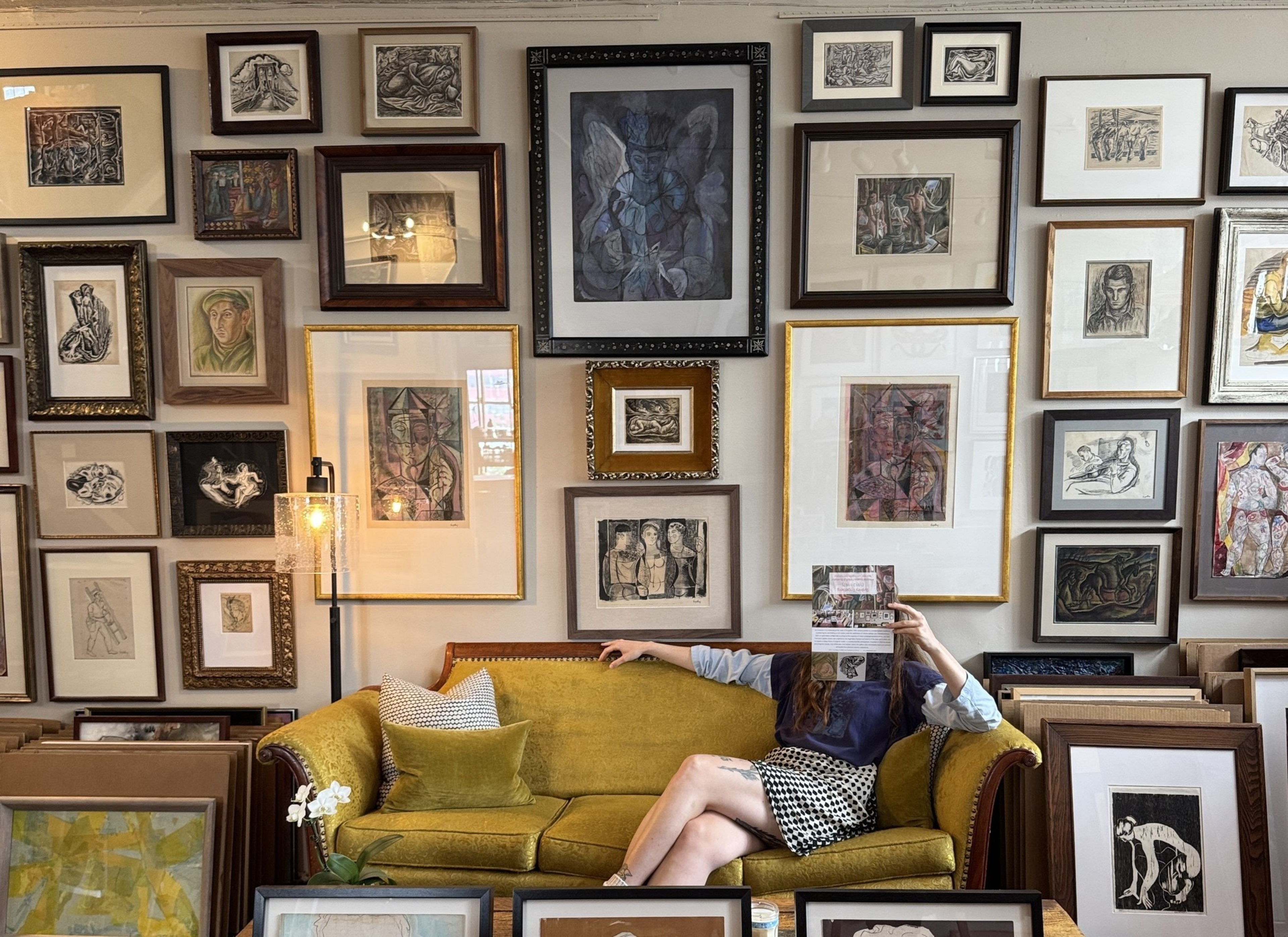 A person lounges on a mustard-yellow couch in a room filled with framed art. They hold a paper in front of their face. Art covers the walls and surrounds the couch.