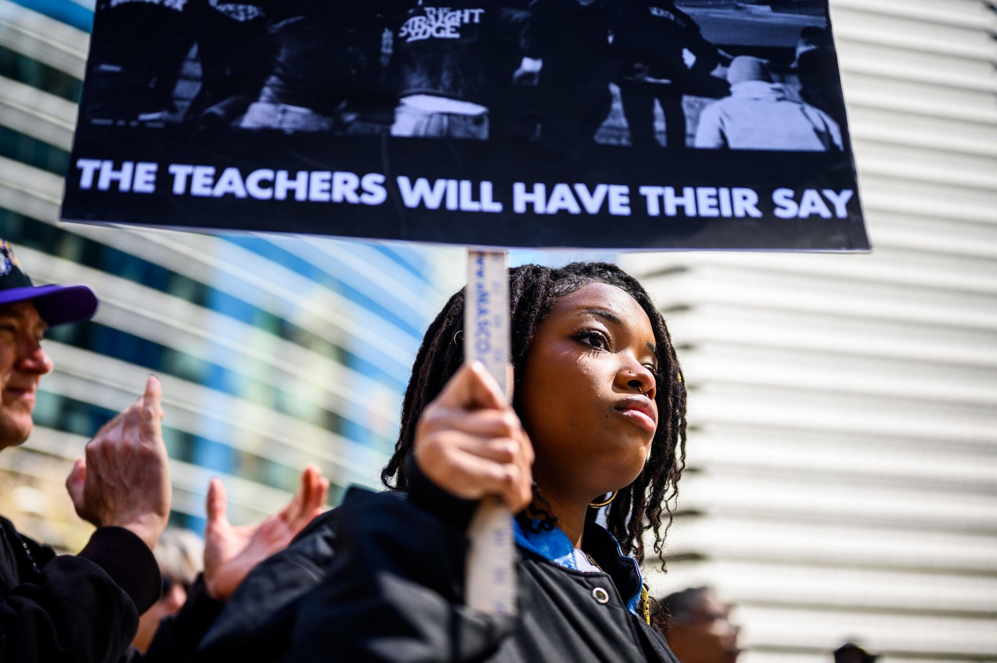 A young woman holds a sign that reads "THE TEACHERS WILL HAVE THEIR SAY" during a protest, with people clapping in the background.