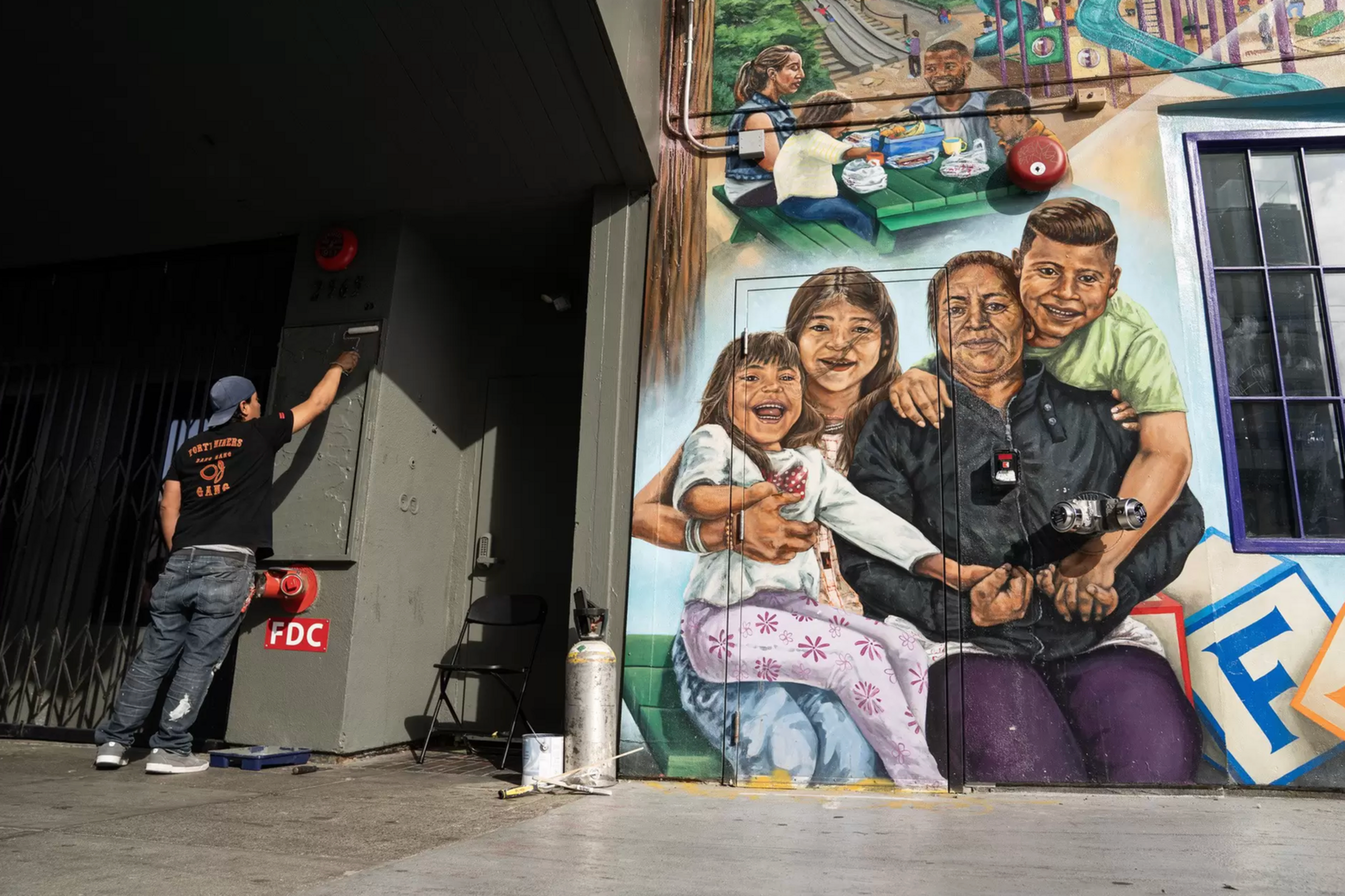 A man is painting a building wall next to a colorful mural of a woman and three children smiling and hugging each other.
