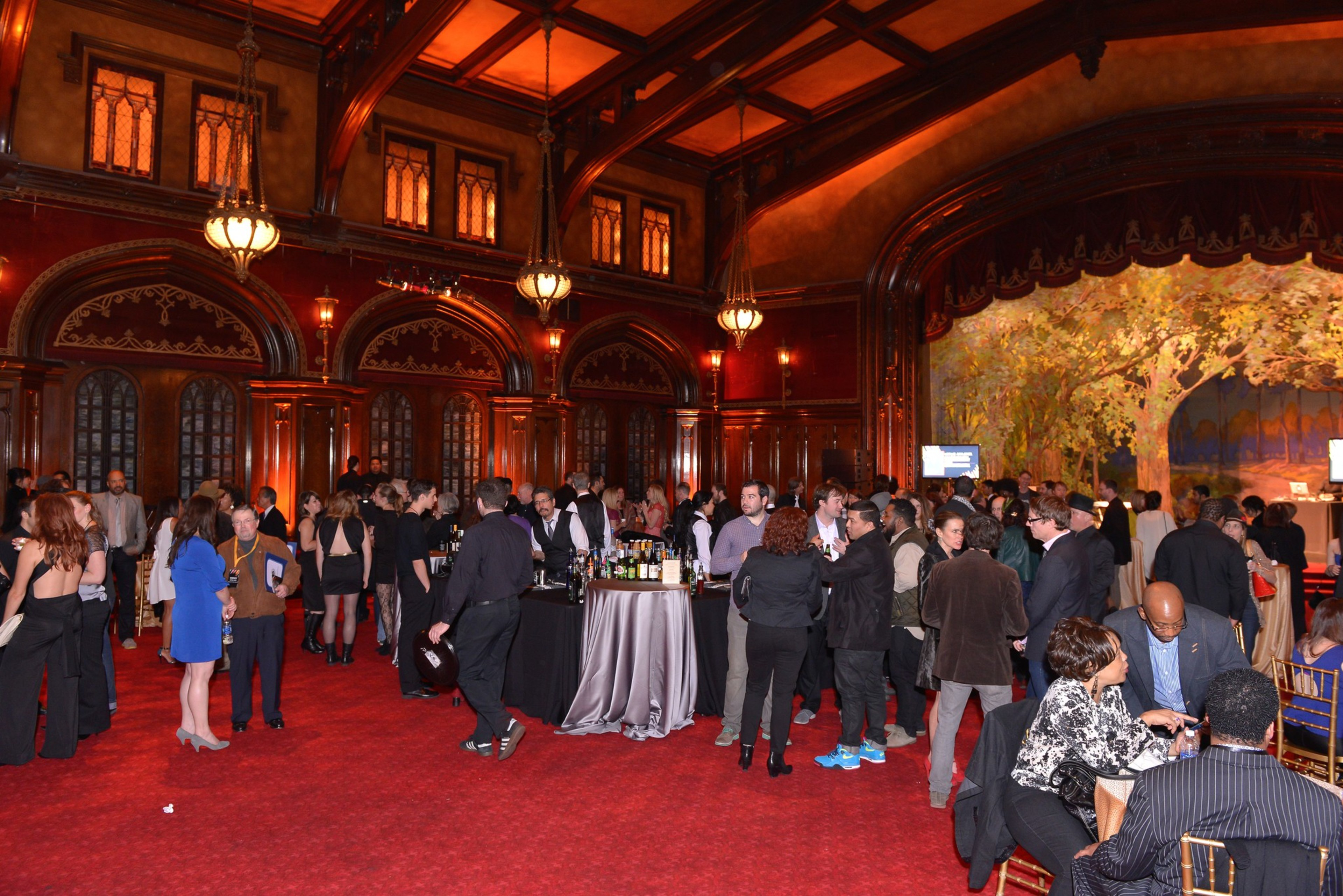 A large, elegant ballroom filled with people socializing around tables, under warm lighting. Ornate wood paneling and a painted backdrop enhance the setting.