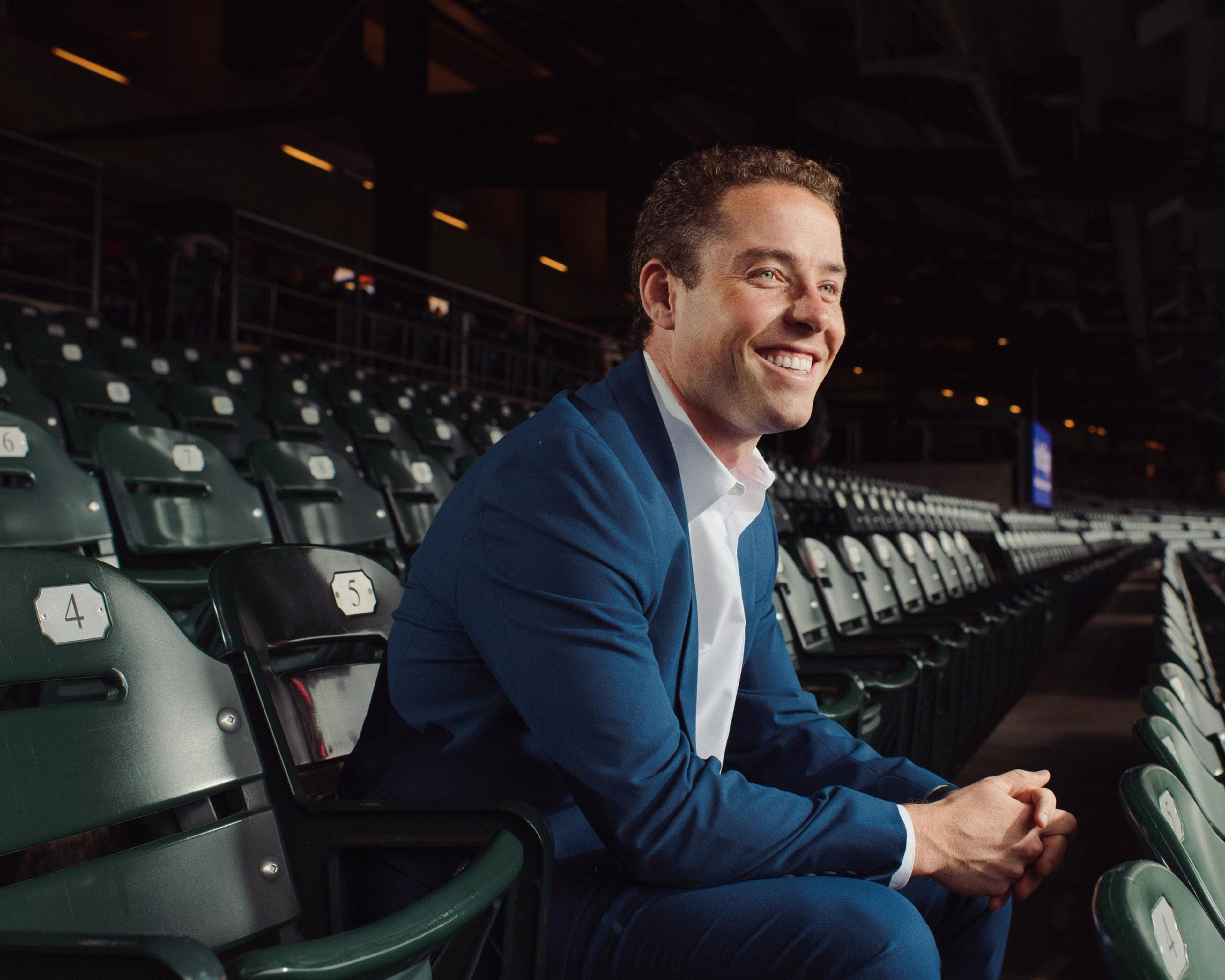 A man in a blue suit smiles while sitting alone in a stadium’s empty green seats, numbered 4 and 5, with dim lighting around him.