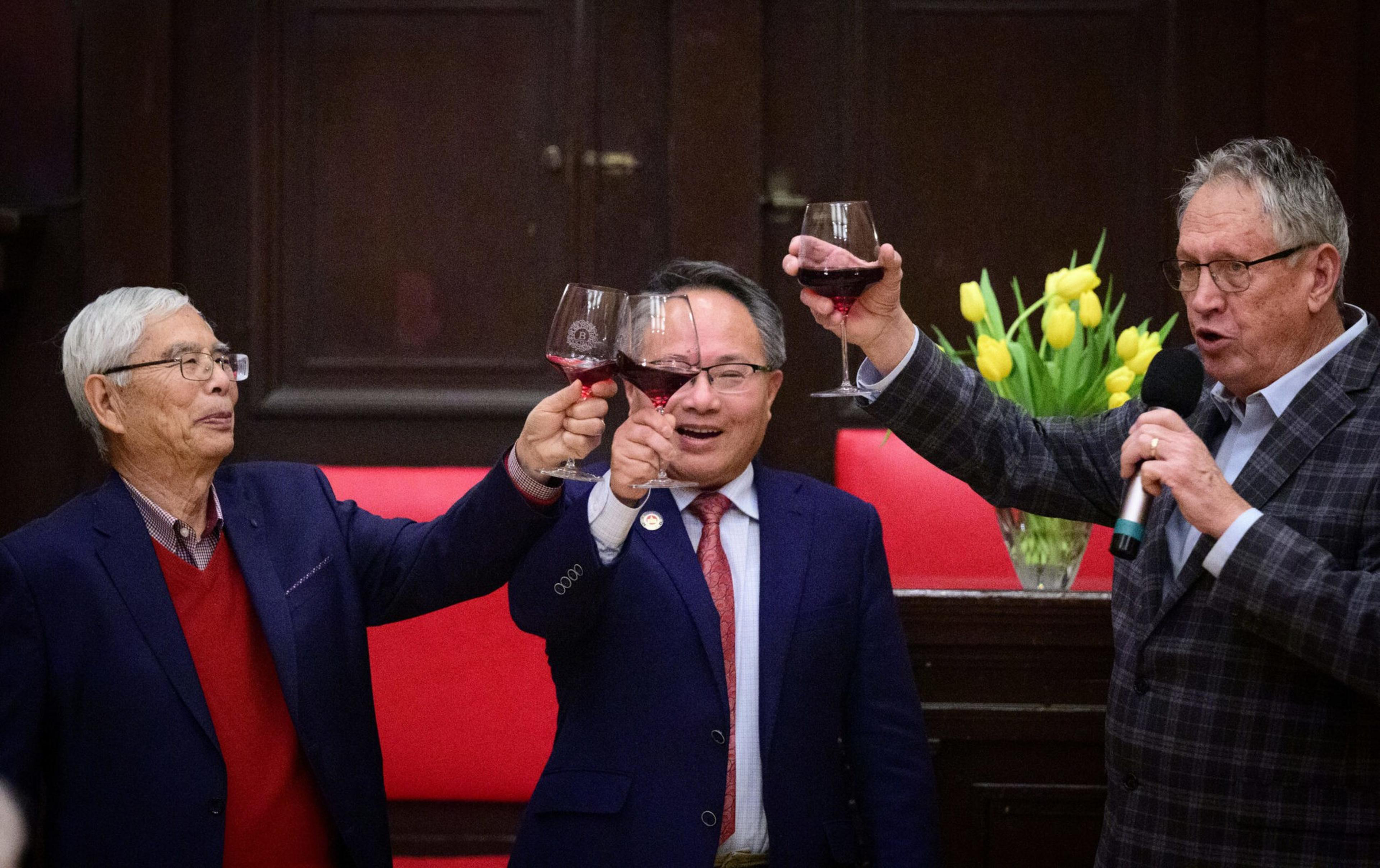 Three elderly men dressed in suits raise glasses of red wine in a toast, while one man speaks into a microphone near a vase of yellow tulips.