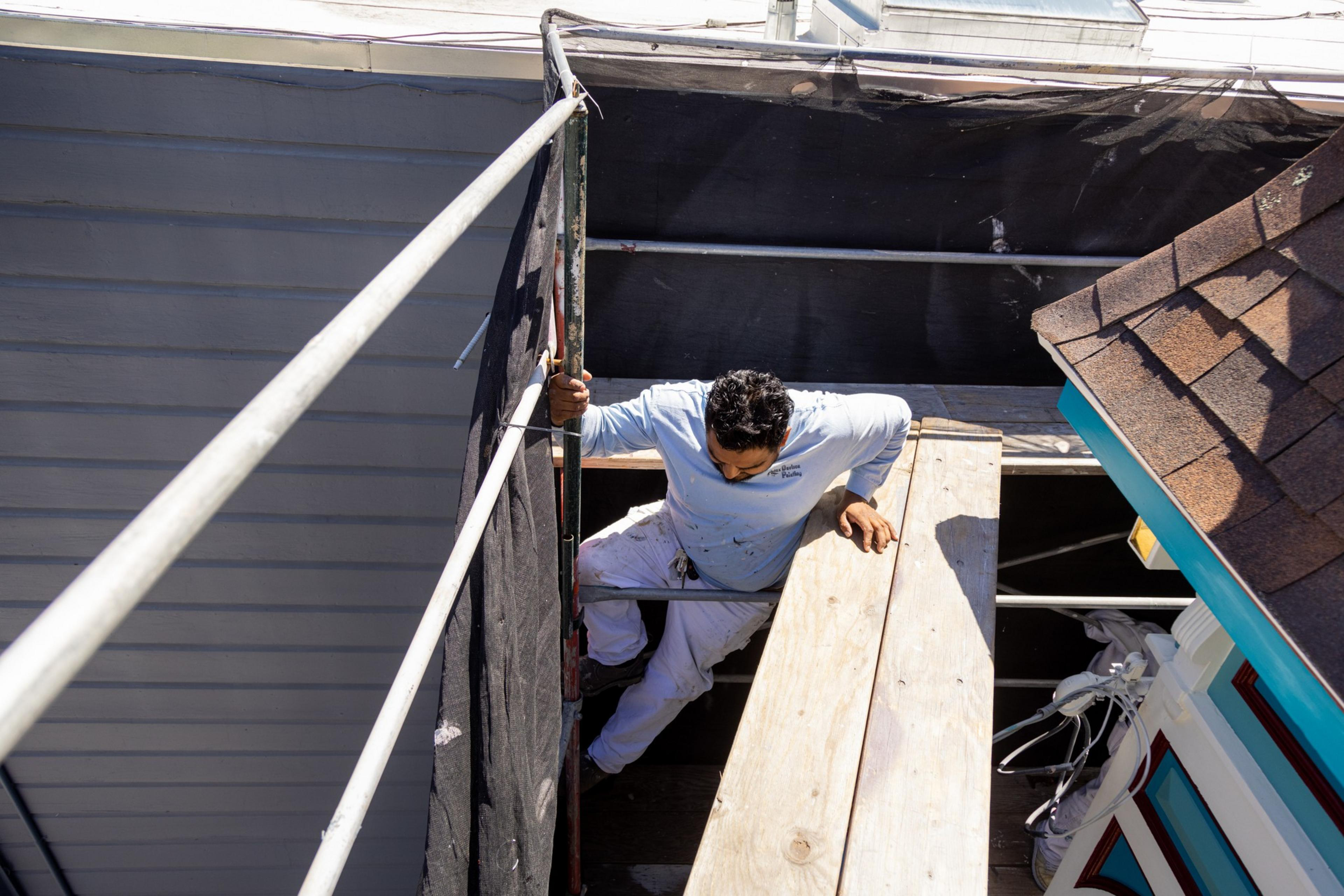 A man in white painter's clothes is climbing out of a dark attic onto a sunlit roof.
