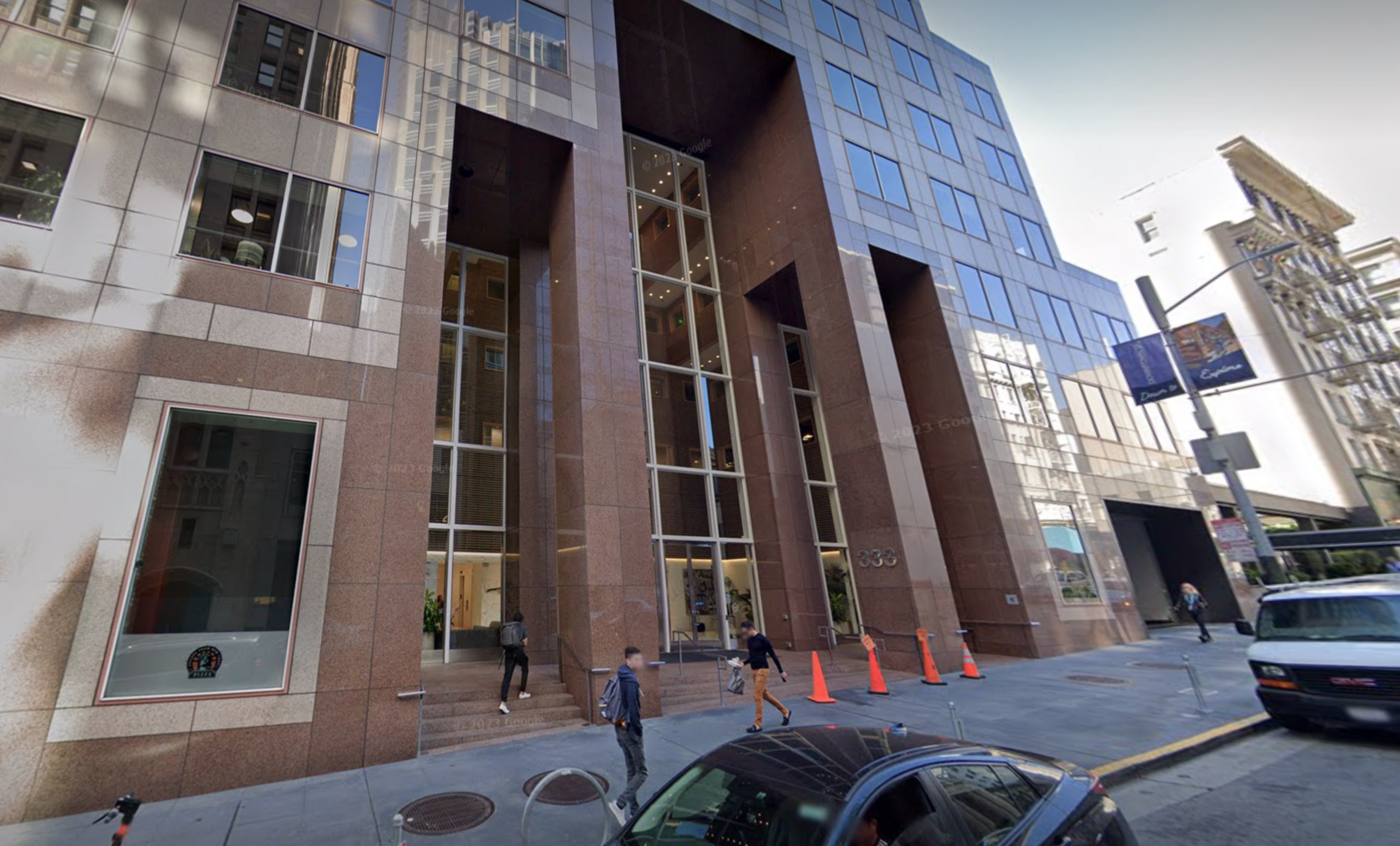 A Google Street View screenshot of a brown corporate building with blurred bystanders walking by