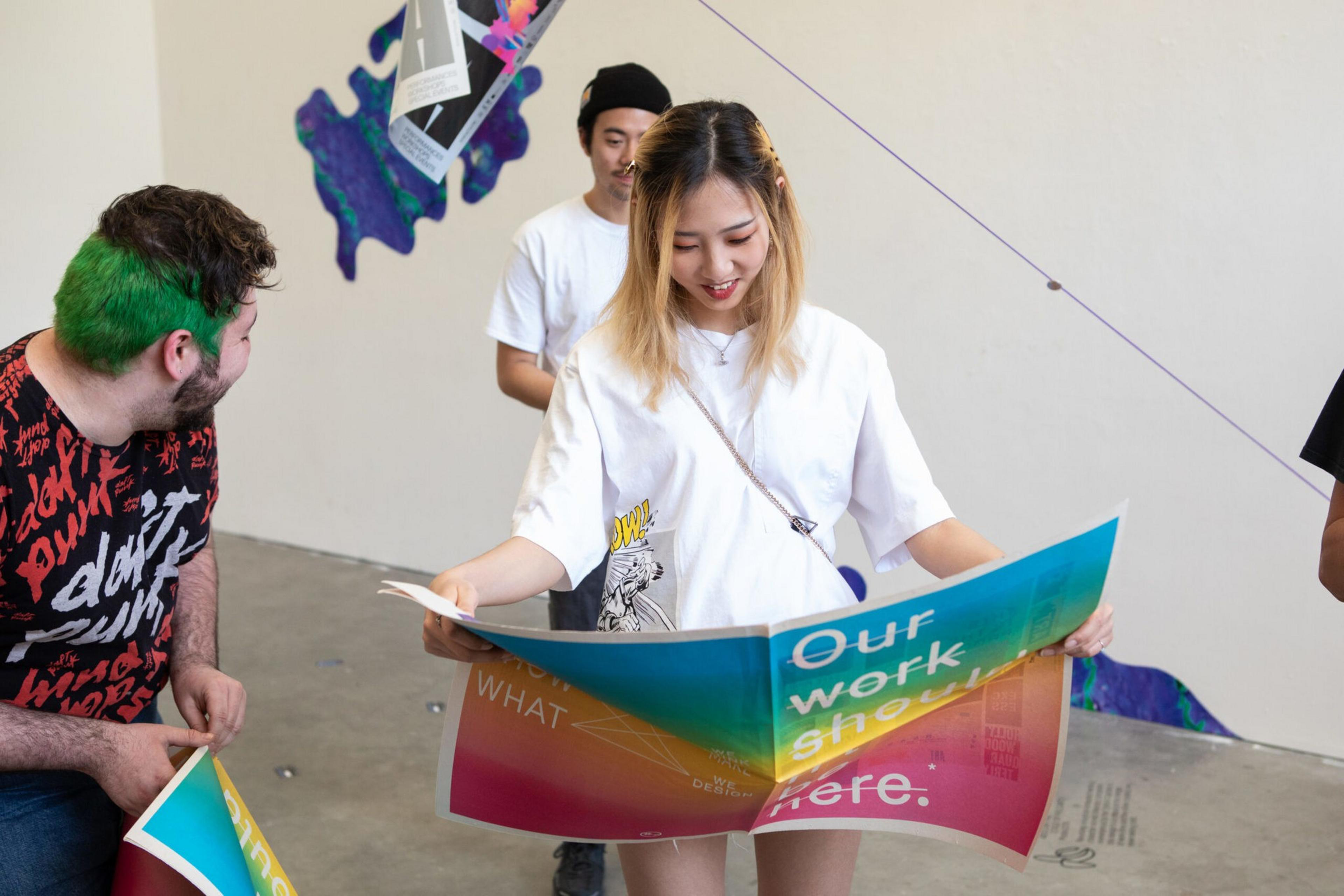 A woman holding and smiling at a colorful large poster, with two men nearby also holding posters, in a bright, casual indoor space.