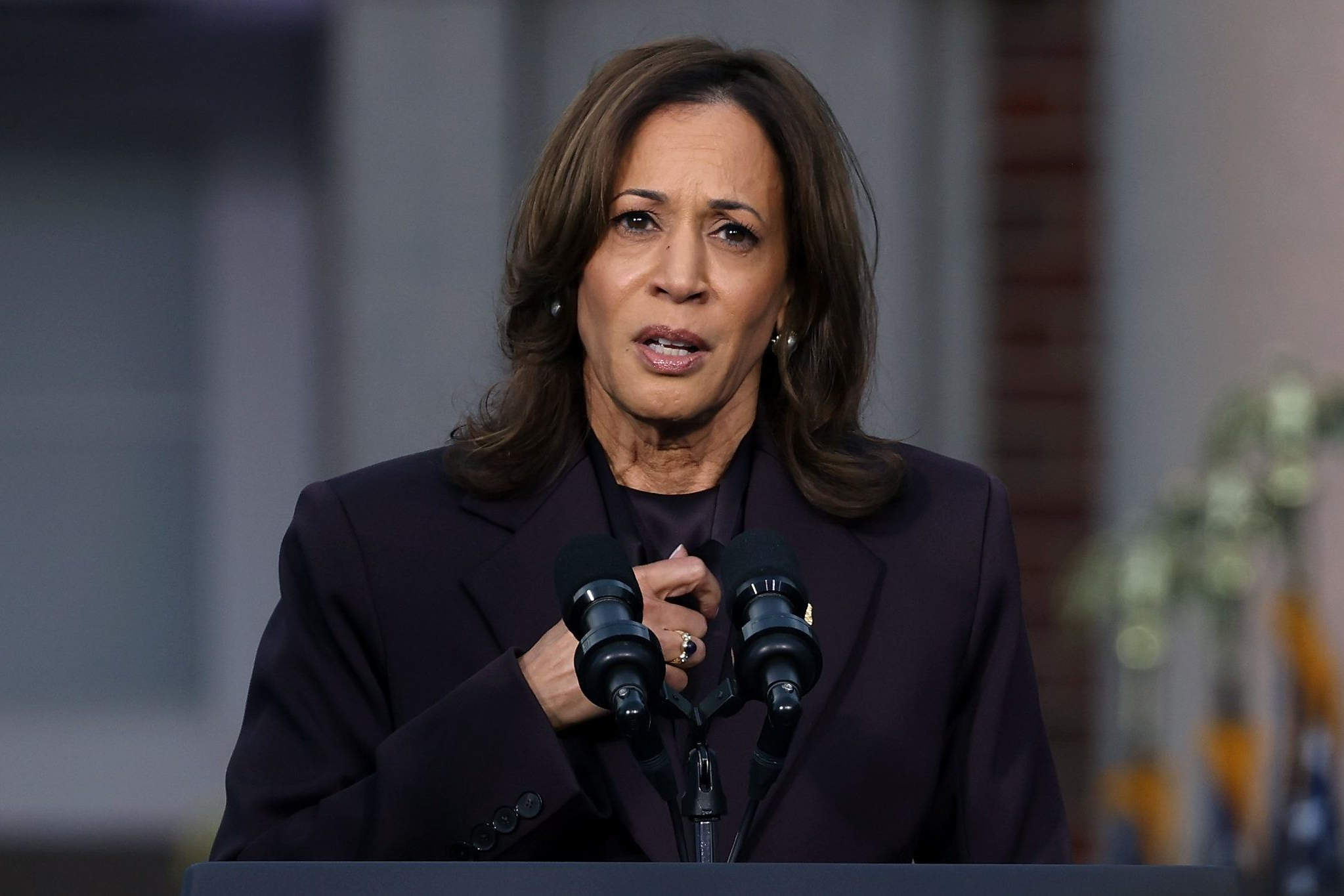 A woman in a dark suit speaks at a podium with "Vice President of the United States" seal. U.S. flags are blurred in the background, and she's speaking into microphones.