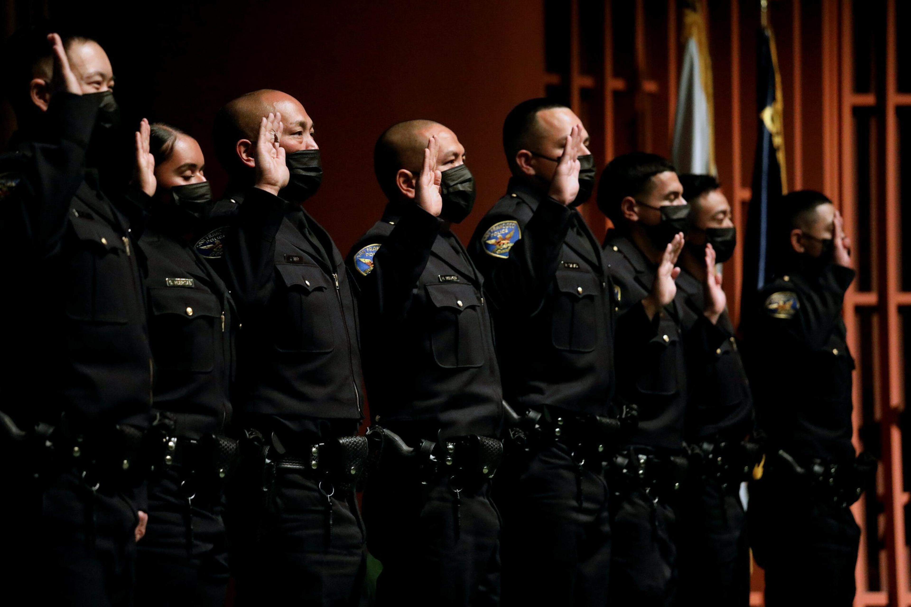 Eight uniformed officers wearing black masks stand side by side with their right hands raised, taking an oath in a formal indoor setting.