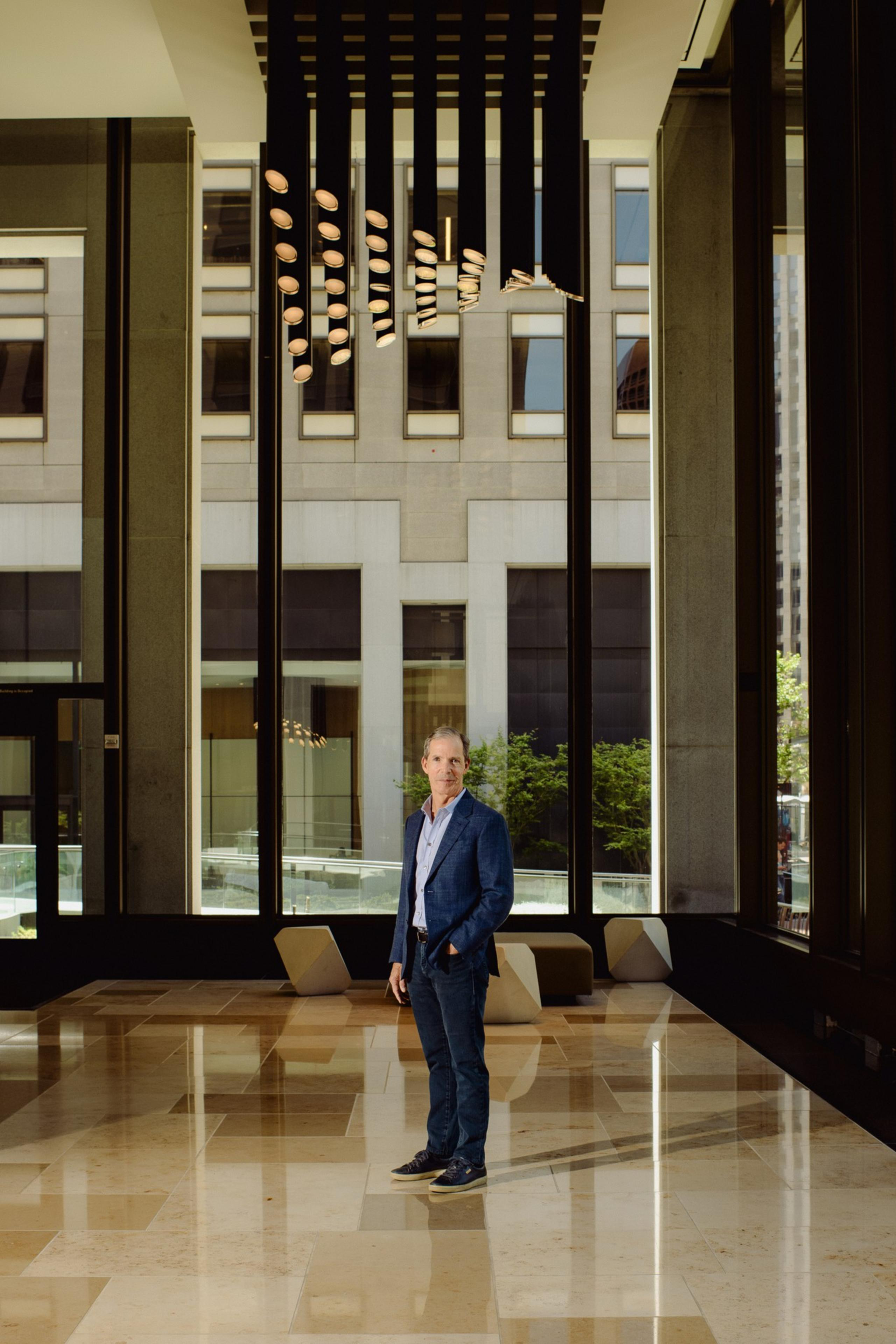 A man in a blue suit stands on a shiny marble floor in a modern building with large windows, letting in natural light. A unique ceiling light fixture is visible.