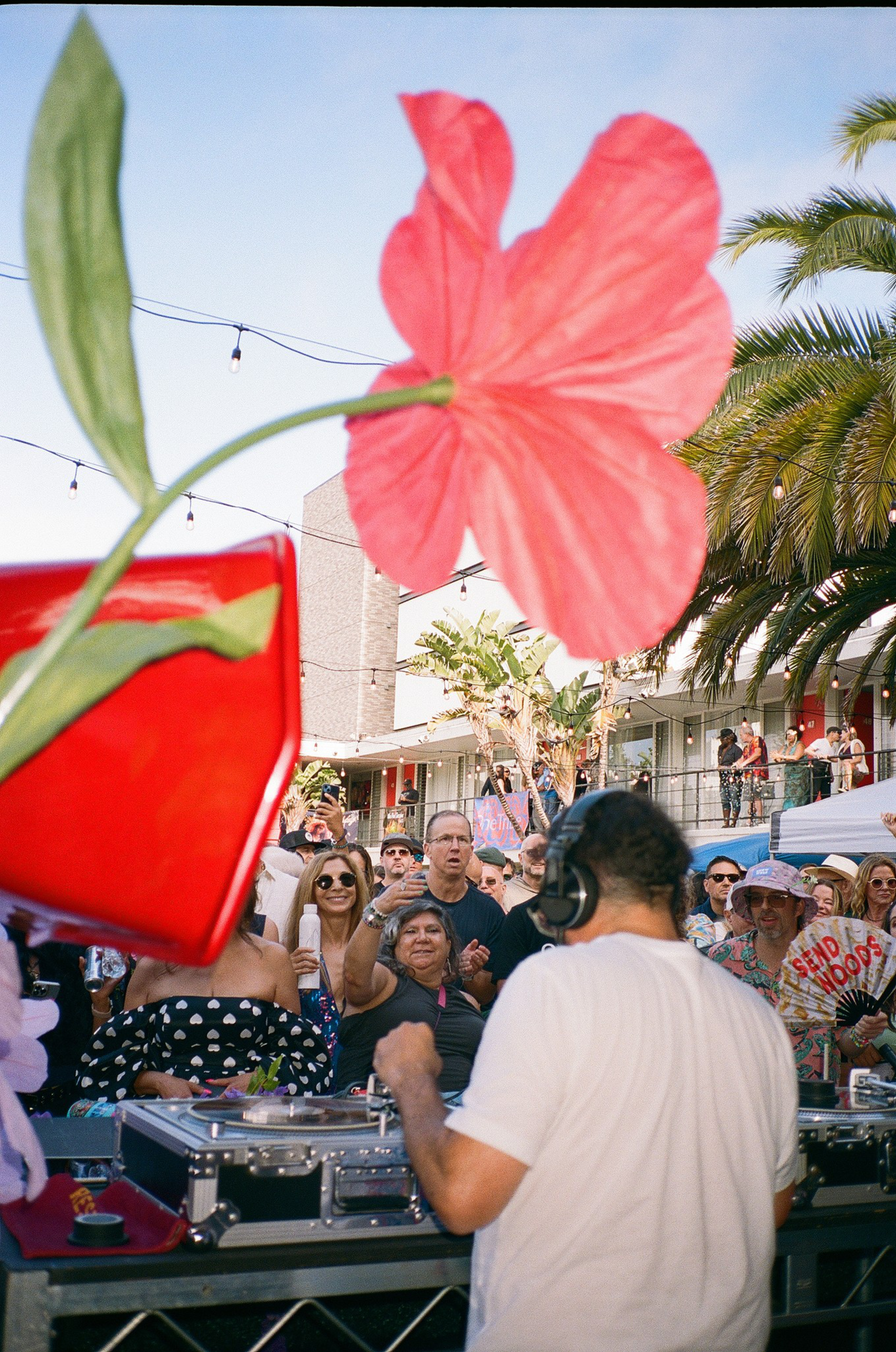 A DJ wearing headphones plays music for a crowd enjoying an outdoor event with palm trees, string lights, and a large red flower decoration in view.