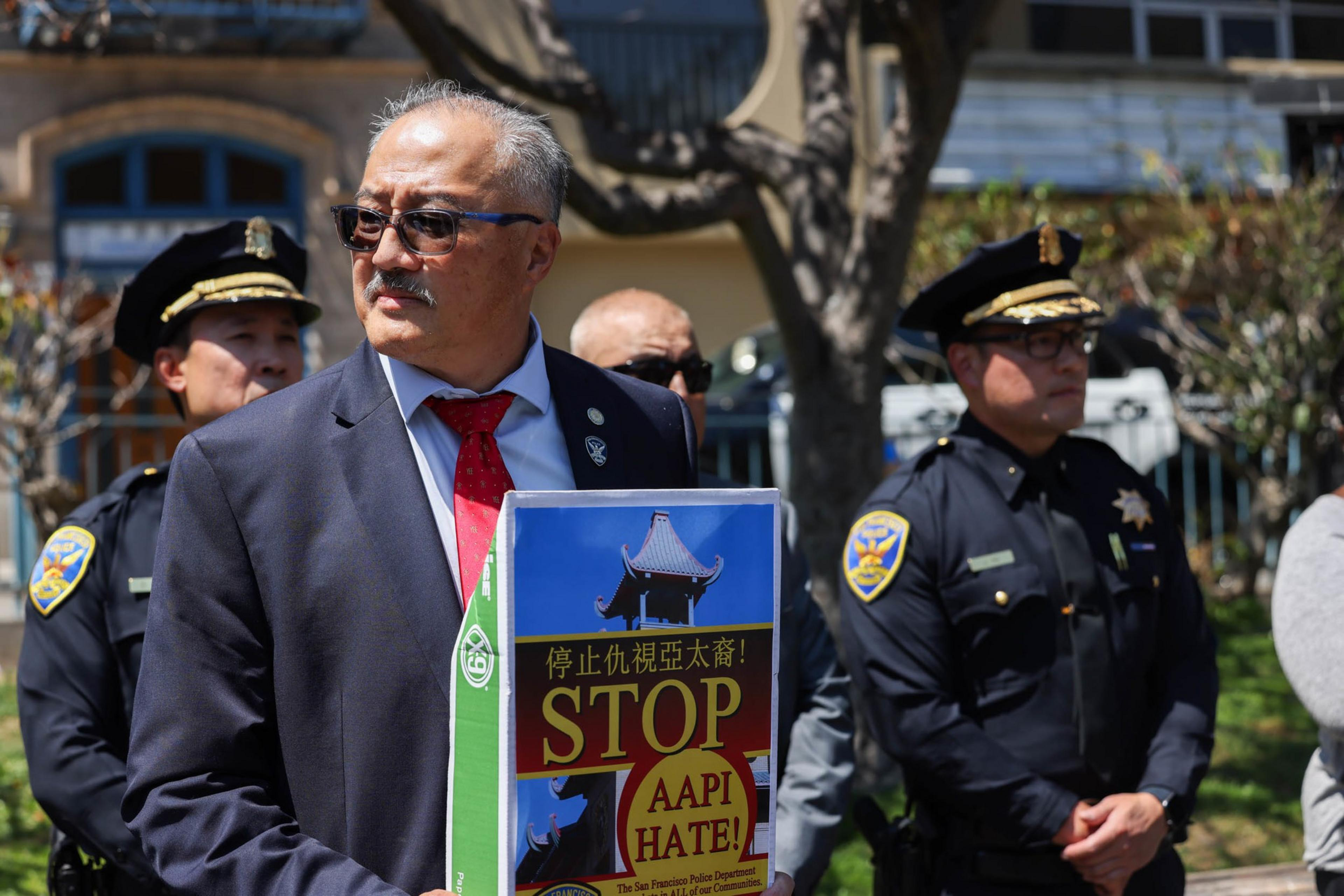 A man in a suit and red tie holds a sign reading "STOP AAPI HATE!" while three police officers stand nearby outdoors.