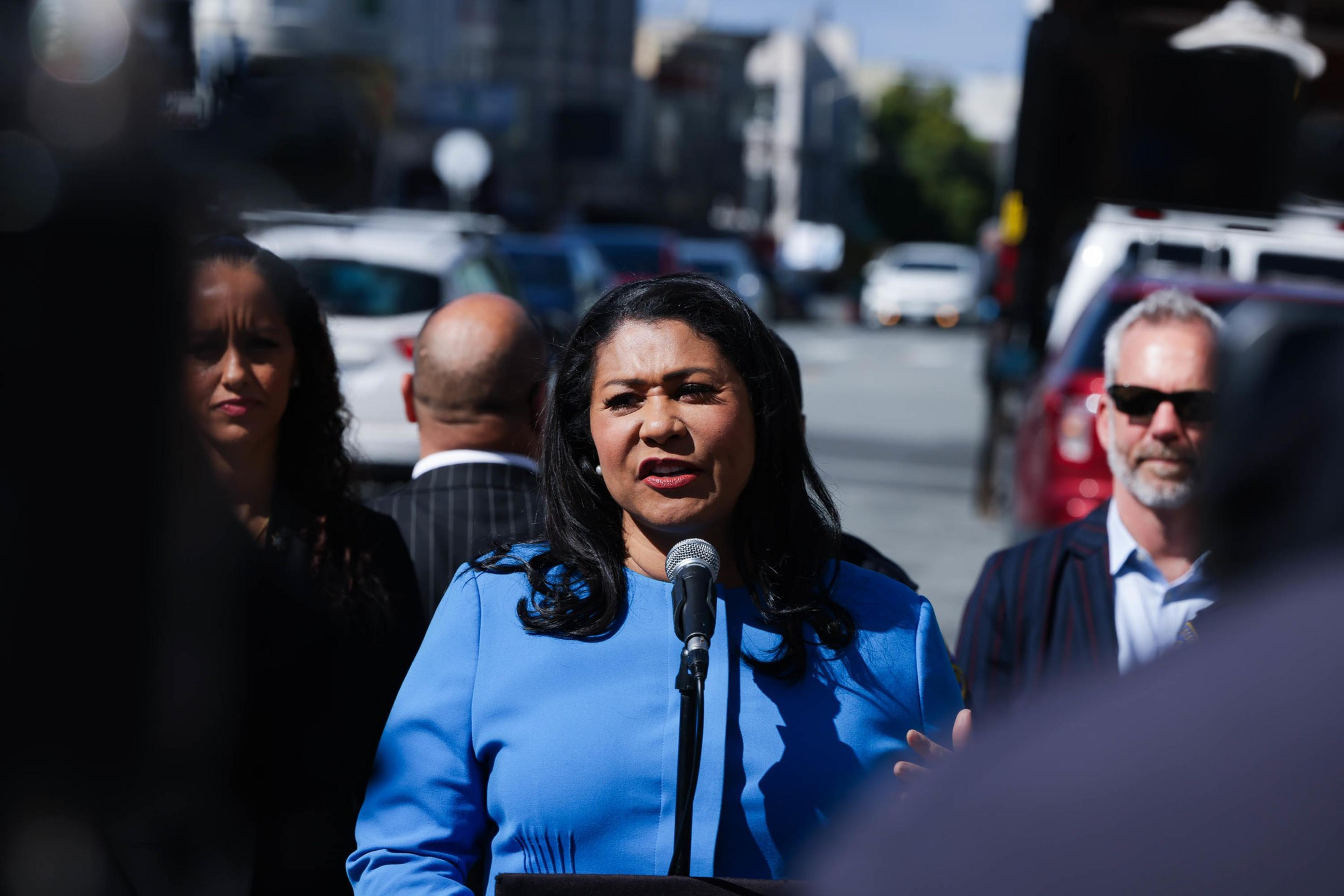 A woman in a blue jacket speaks into a microphone with blurred figures and vehicles in the background.