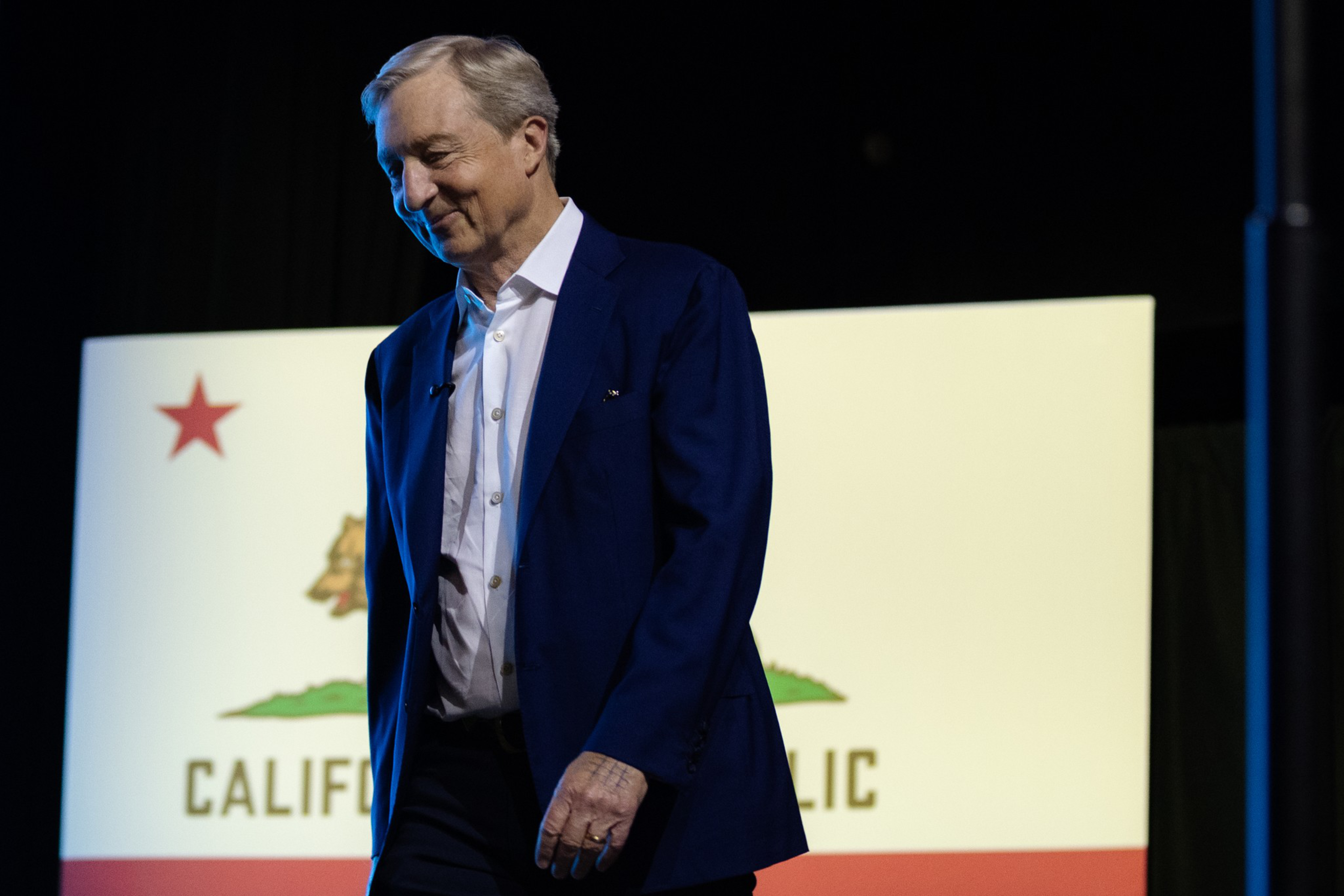 A man in a dark blue suit and white shirt stands in front of a California state flag, smiling slightly with his head tilted down.