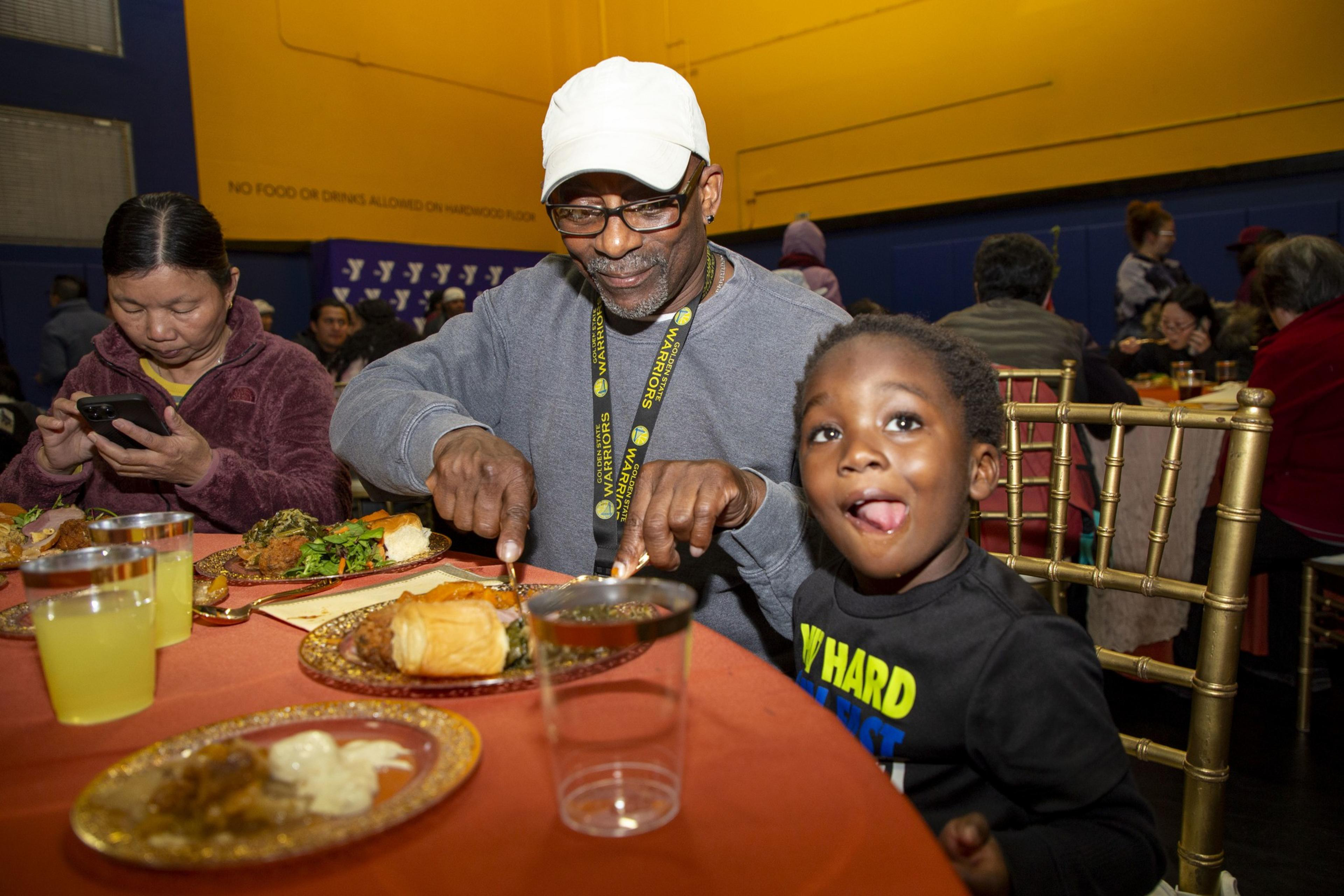 A man in a white cap and glasses smiles while cutting food on his plate. A child beside him playfully sticks out their tongue, sitting at a decorated table.