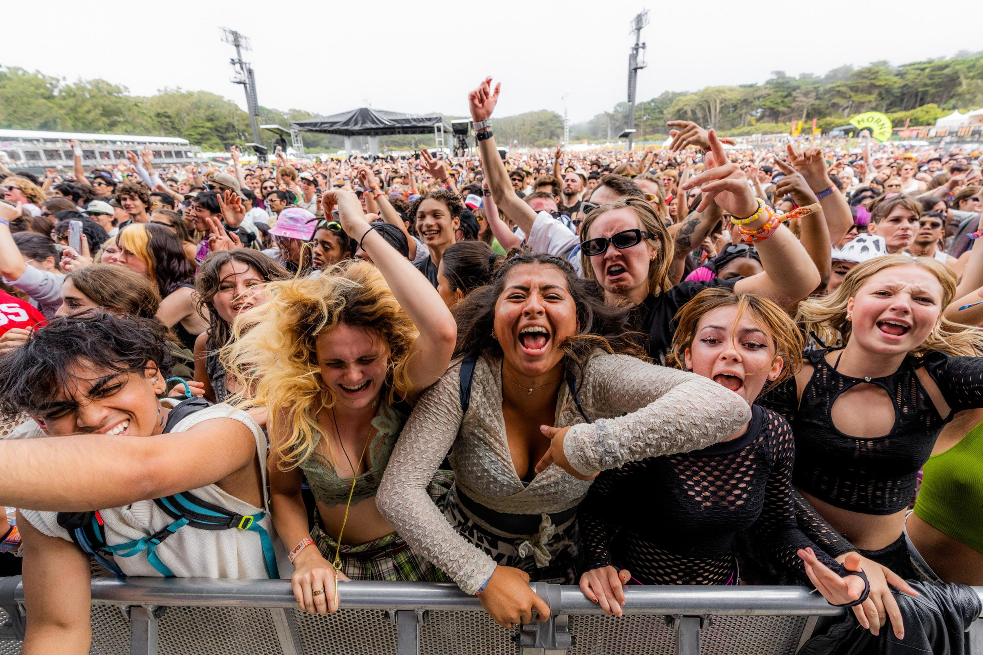 A large joyous crowd gathers at a music festival, with excited fans at the front cheering, laughing, and singing, displaying a lively and energetic atmosphere.