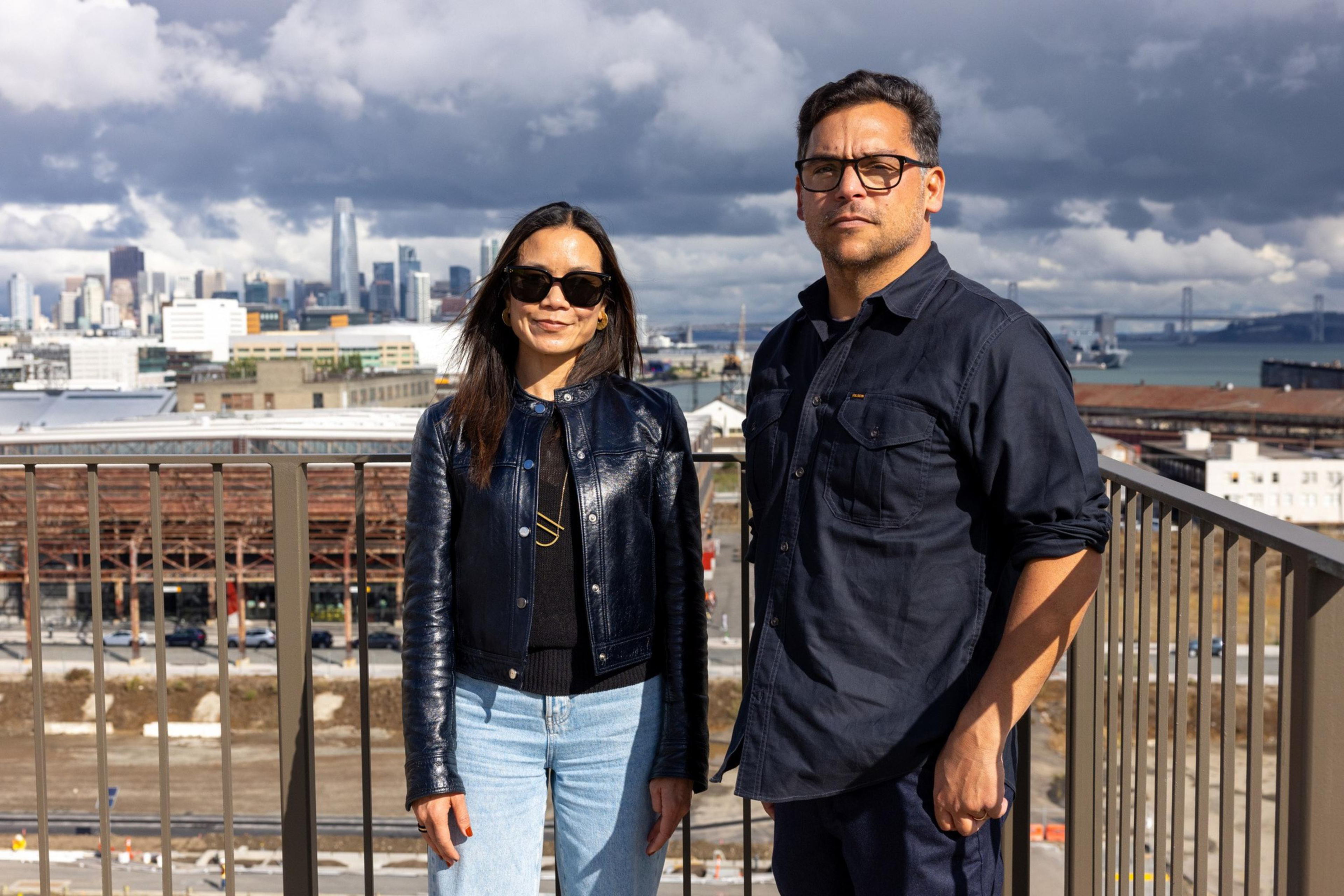 A woman and a man stand on a rooftop with a city skyline and water in the background, both wearing sunglasses and dark tops.