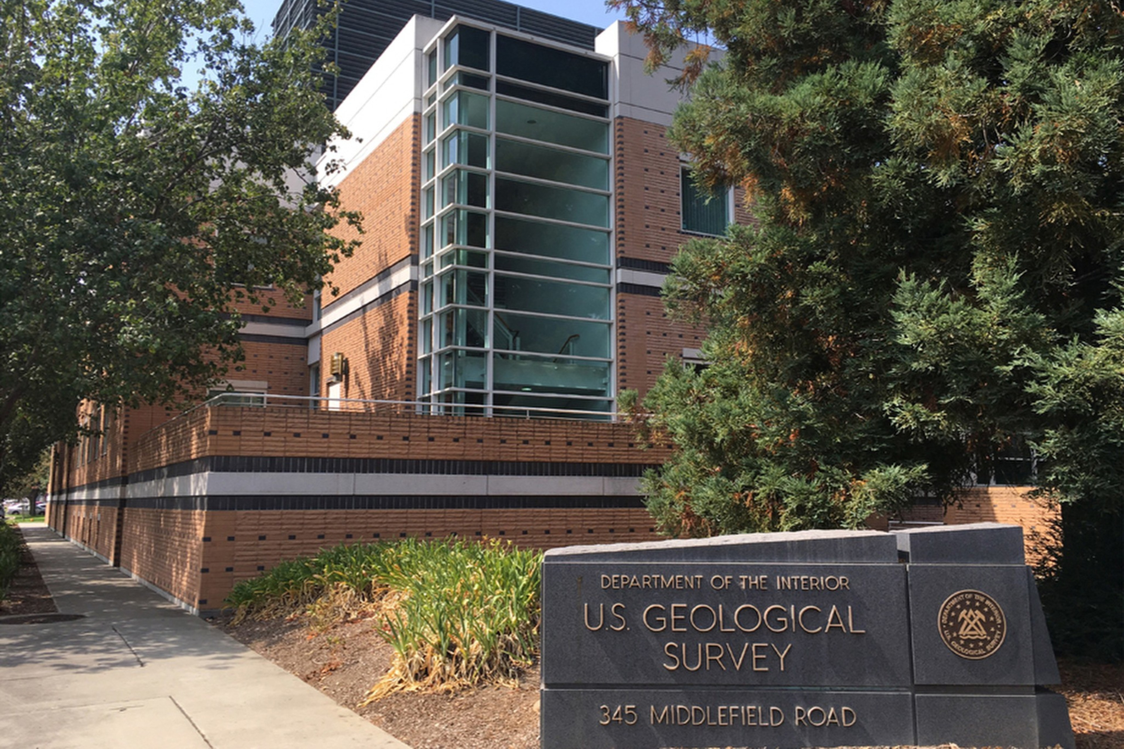 The image shows a brick building with large glass windows, surrounded by trees. A sign in front reads "U.S. Geological Survey, 345 Middlefield Road."