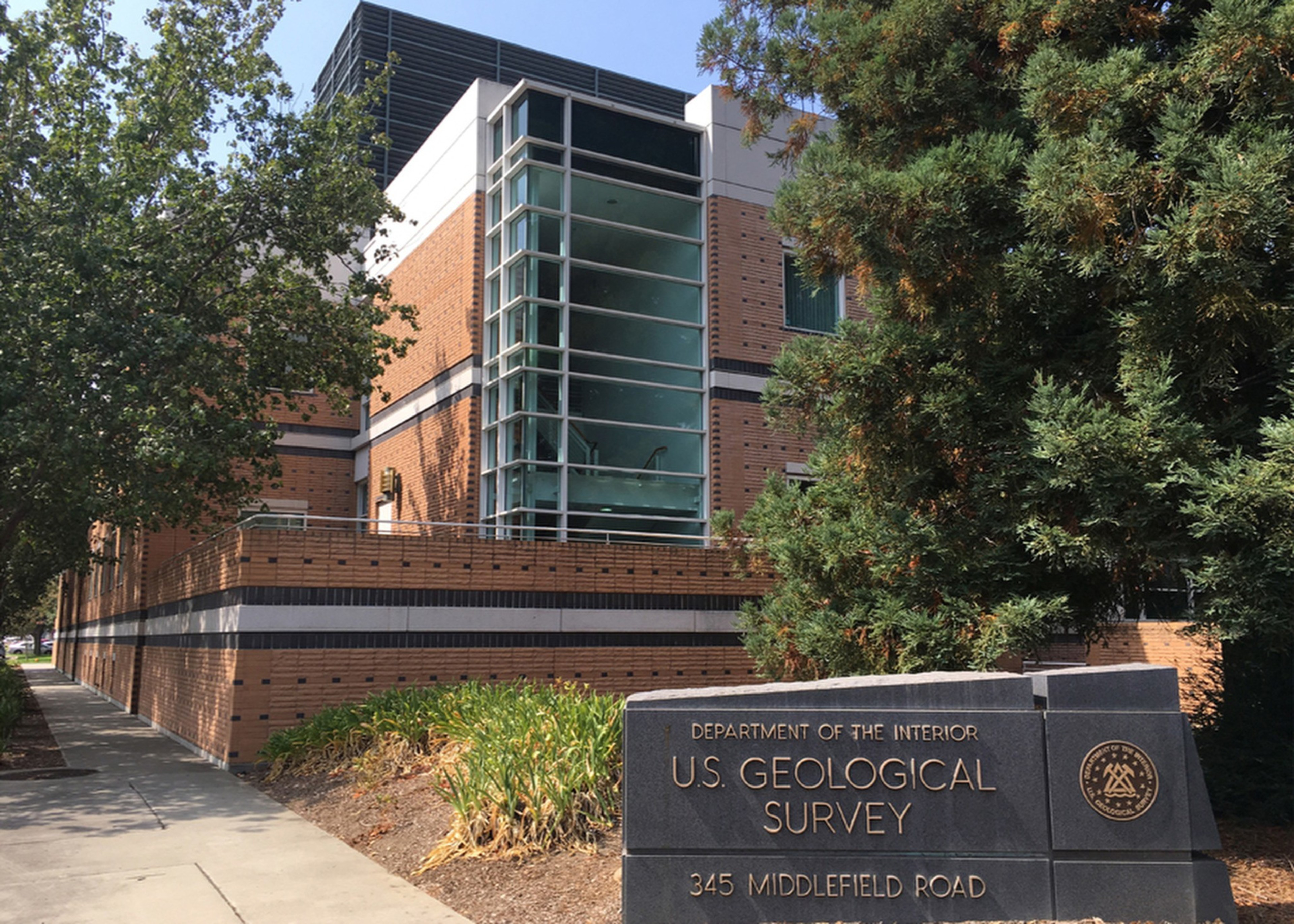 The image shows a brick building with large glass windows, surrounded by trees. A sign in front reads "U.S. Geological Survey, 345 Middlefield Road."