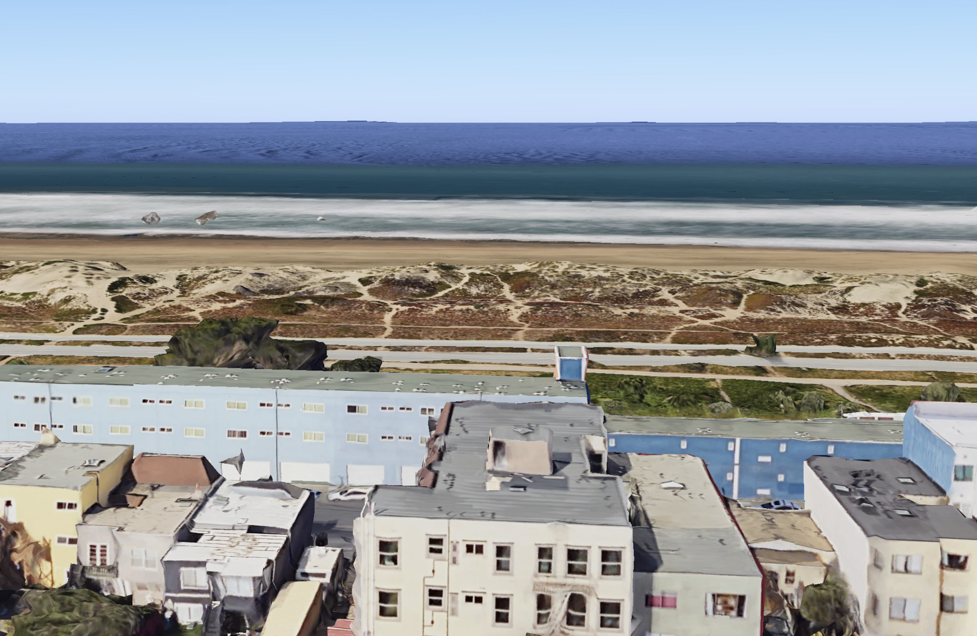 The image features a coastal scene with a clear blue ocean, sandy beach, grassy dunes, and buildings in the foreground. The sky is clear and blue.