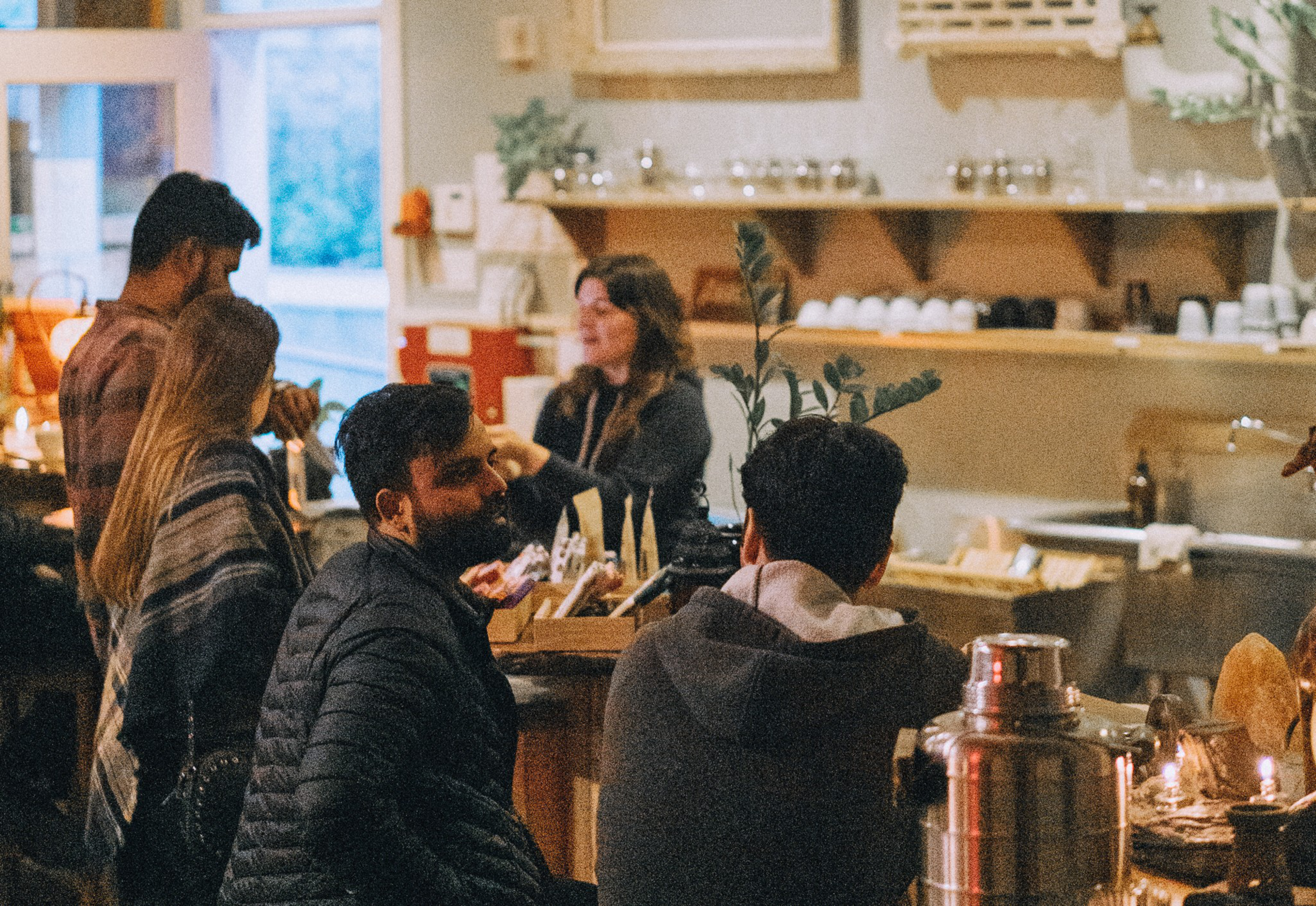 Four people are chatting in a cozy café. The scene includes warm lighting, shelves with cups, plants, and a barista in the background.