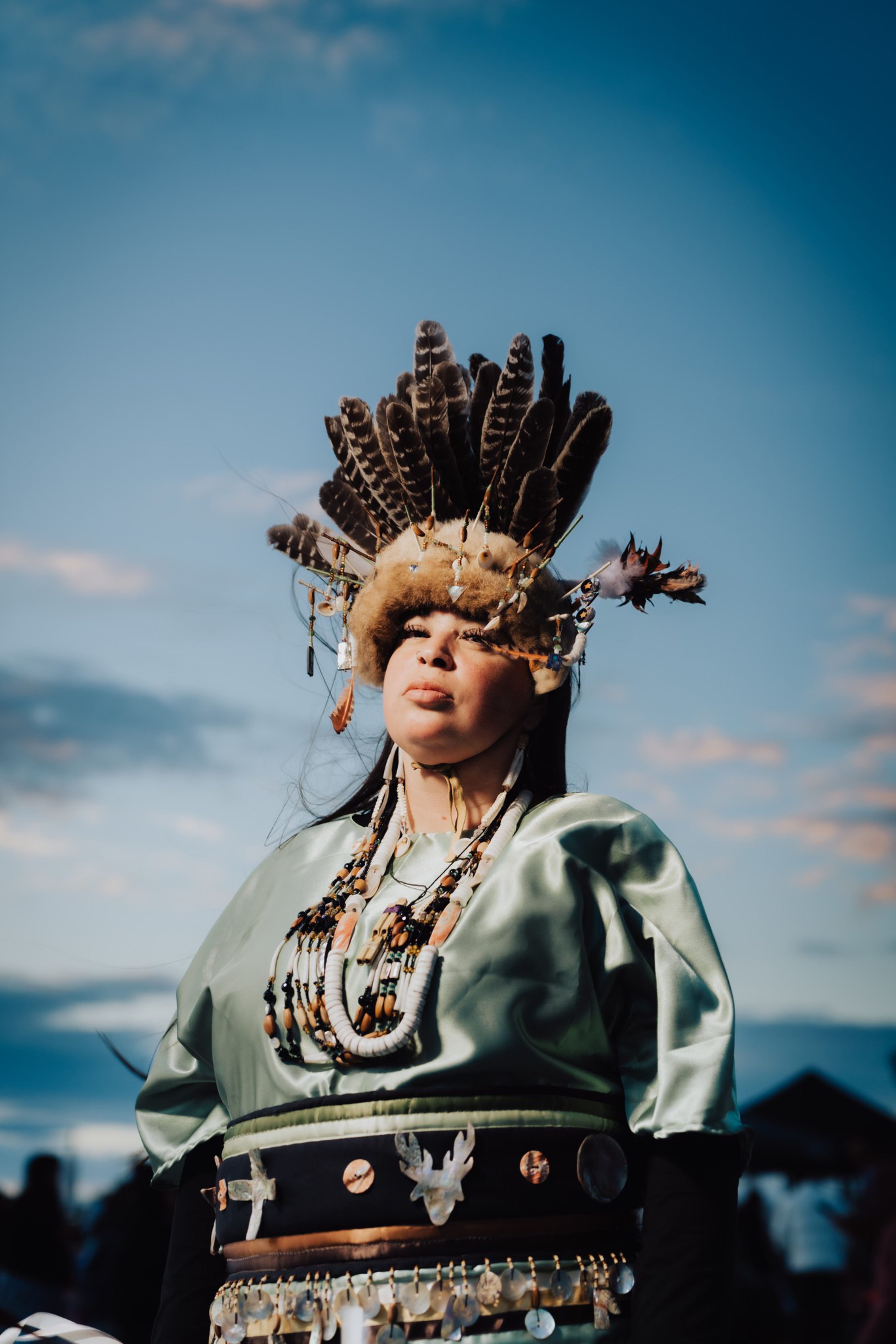 A person wears traditional regalia with a large feathered headdress, beaded necklaces, and a decorated belt, standing against a cloudy blue sky.