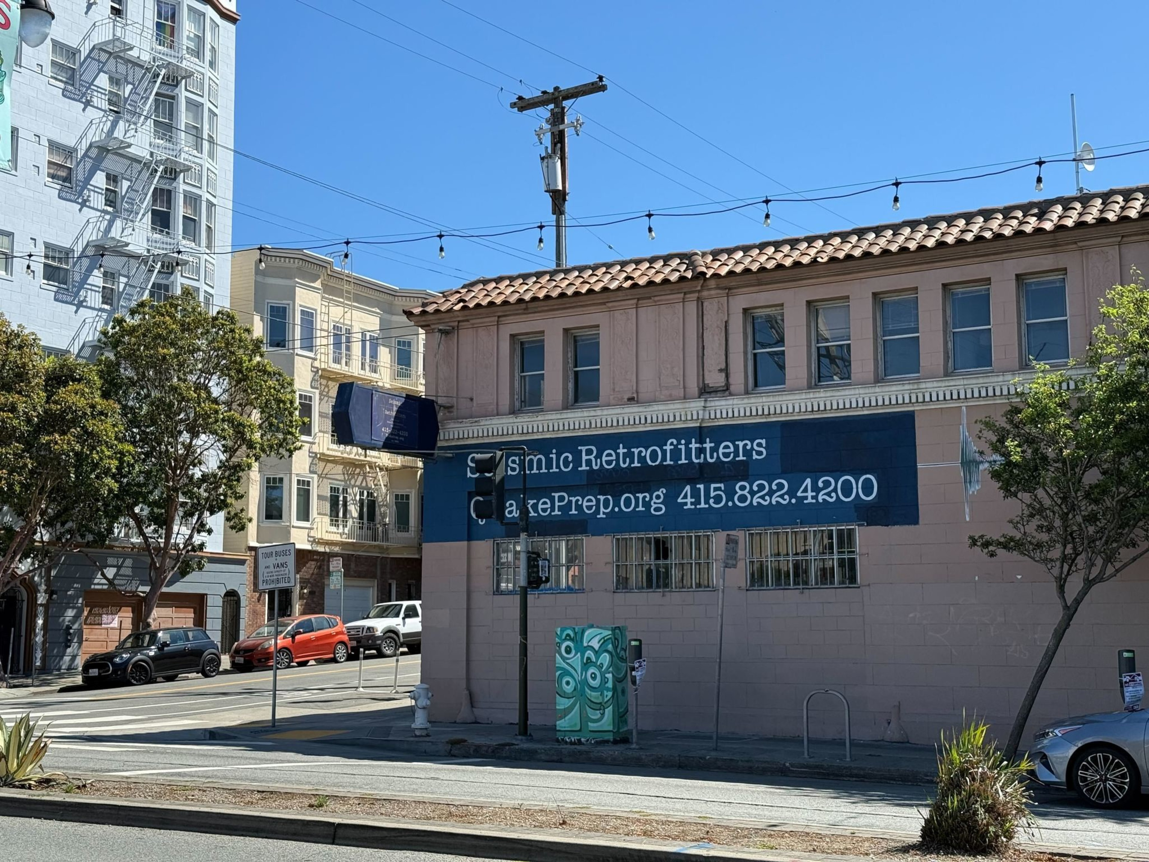 A two-story building has a blue sign reading "Seismic Retrofitters" and "quakePrep.org 415.822.4200," with trees, parked cars, and string lights above.