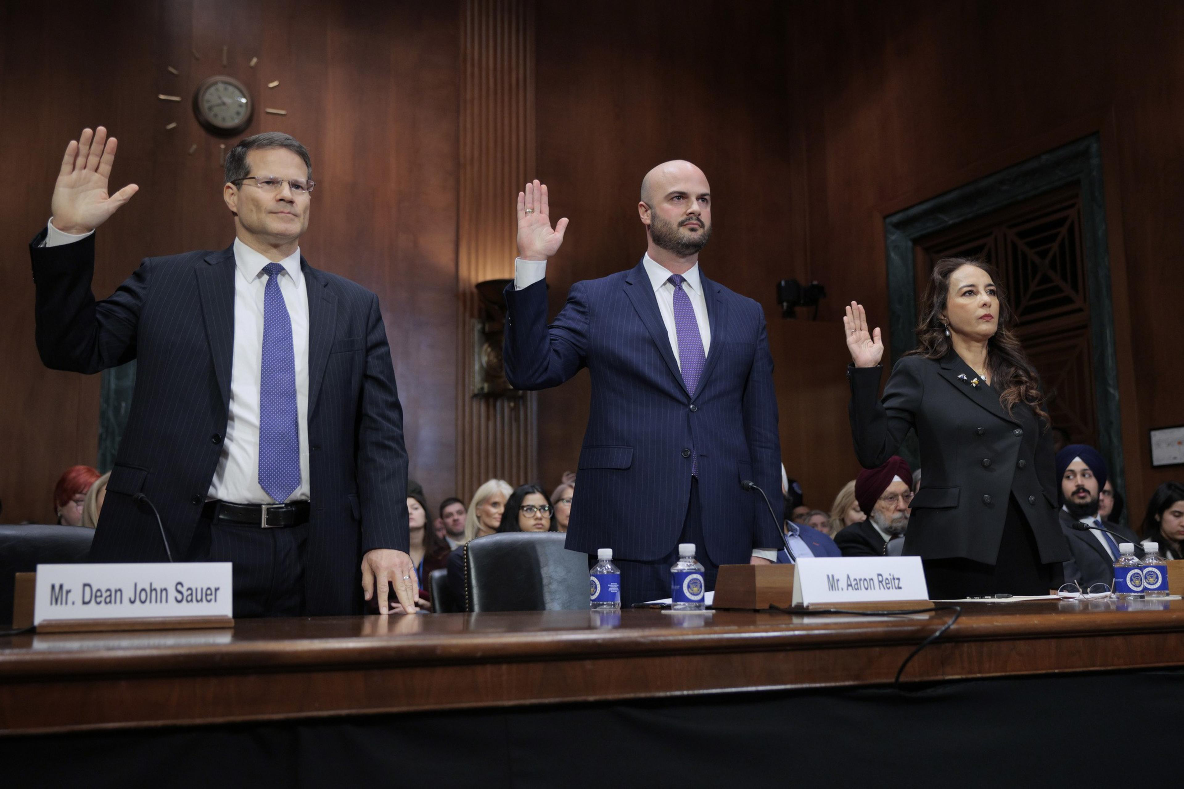 Three people in formal attire stand with raised right hands, likely during a hearing or swearing-in, in a wood-paneled room with an audience seated behind them.