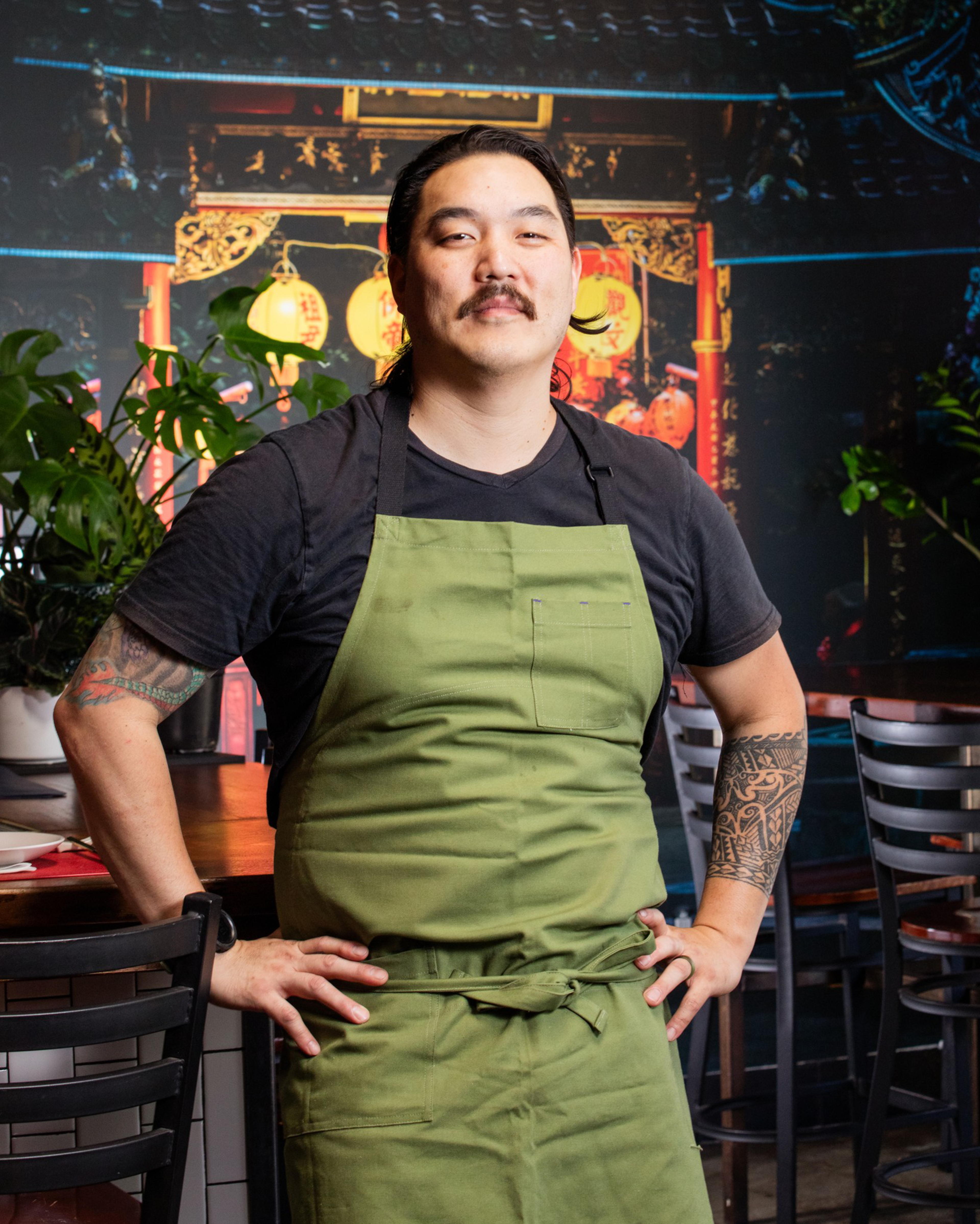 A man with black hair and a mustache wears and apron and black t-shirt leans against a bar with his handson his hips.