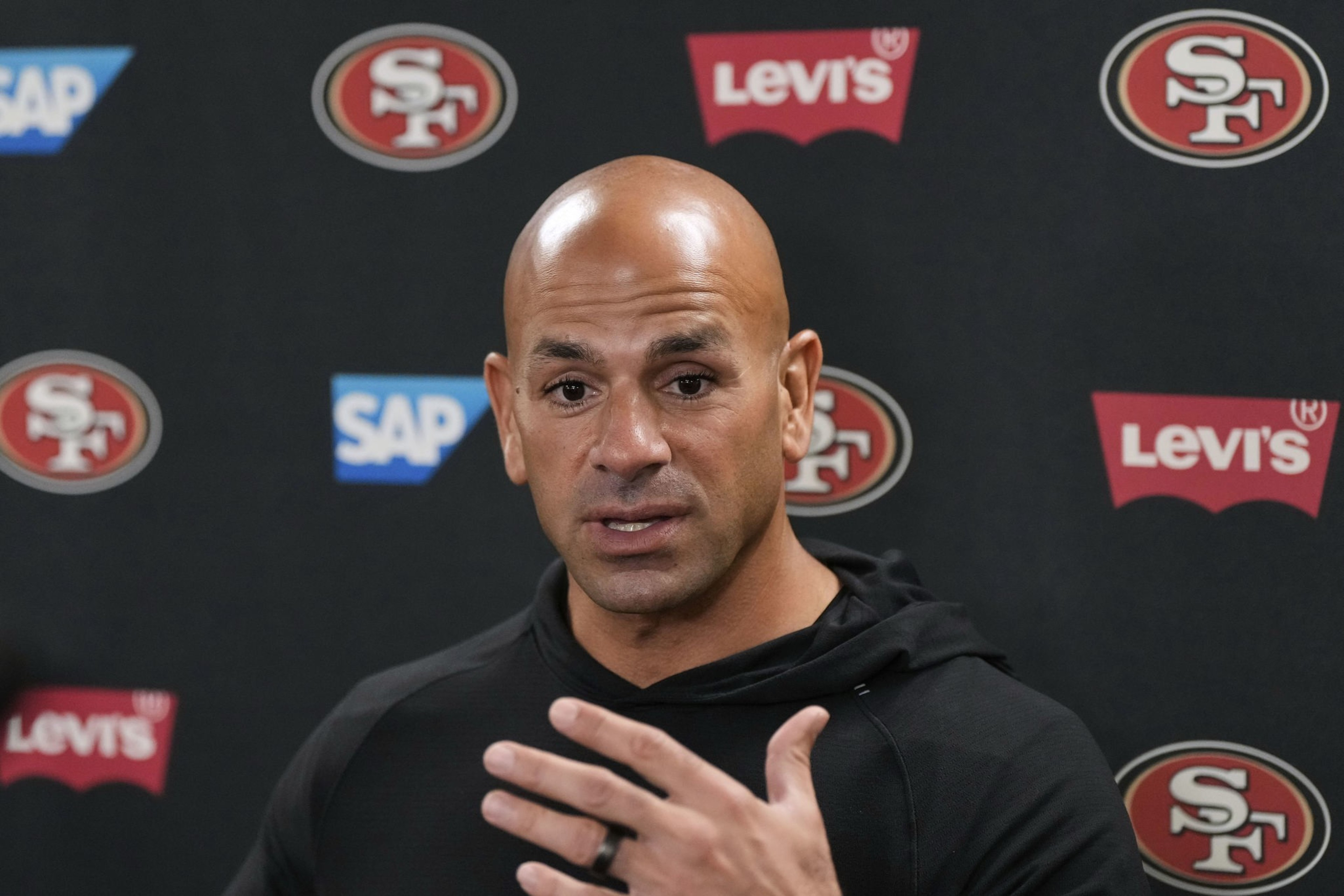 A bald man in a black hoodie speaks, gesturing with his hand. Behind him are logos for the San Francisco 49ers, SAP, and Levi's on a black background.