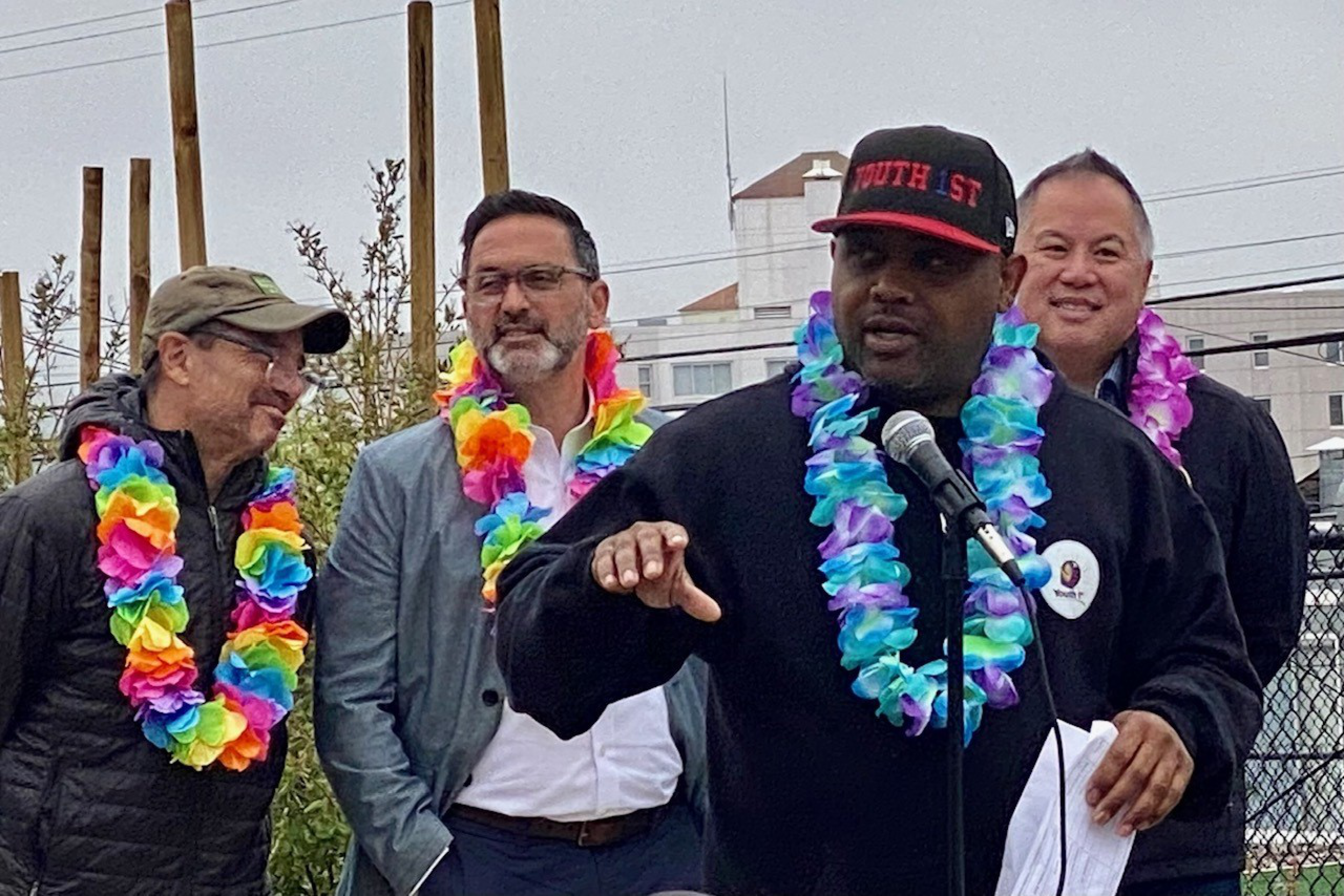 Four men wearing brightly colored flower leis, one speaking into a microphone while holding papers, outdoors with buildings and plants in the background.