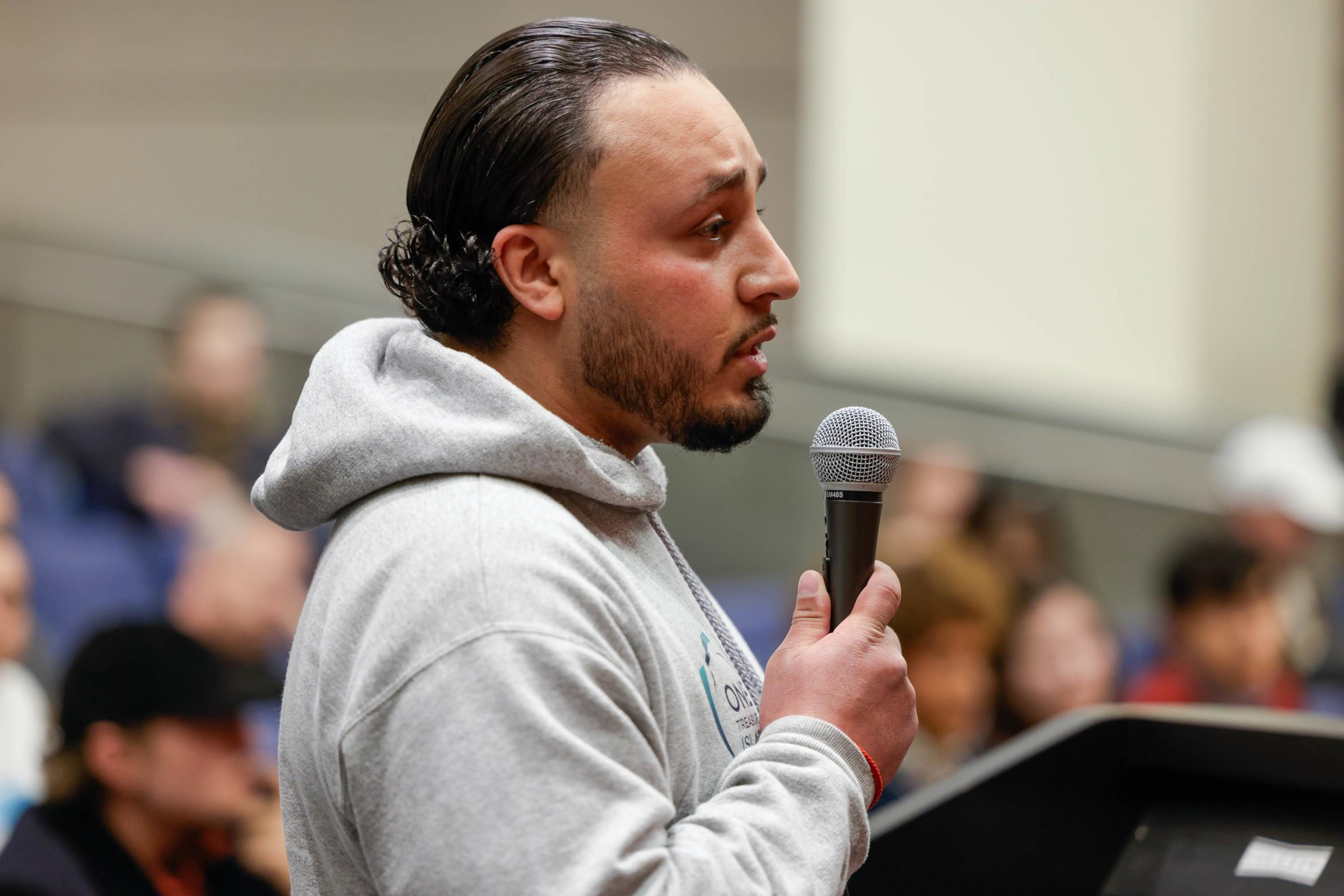 A man with long black hair and a gray hoodie speaks at a podium, seen in a profile side-angle.