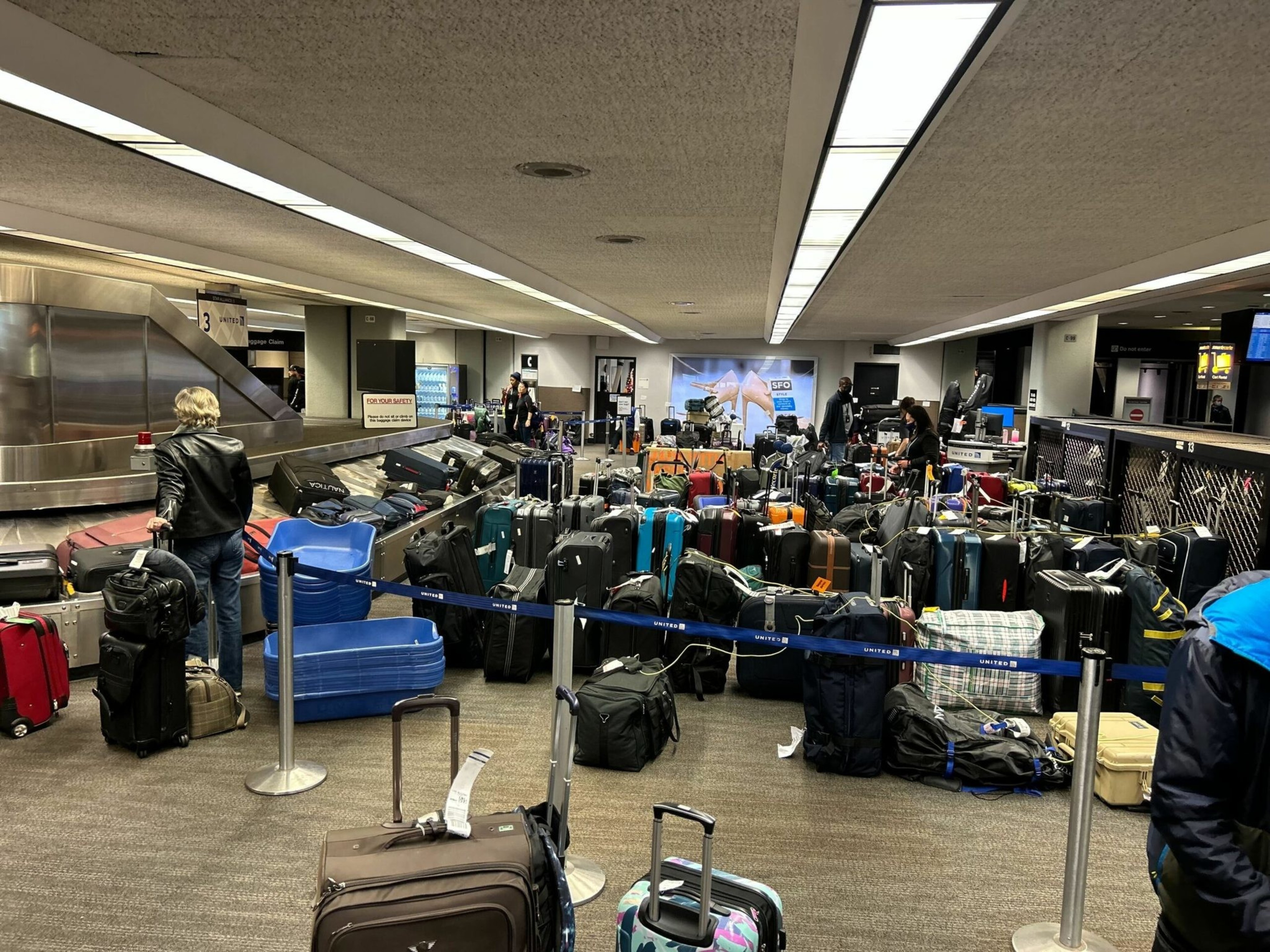 Numerous suitcases and bags are piled inside a baggage claim area at an airport, with a few travelers waiting nearby.