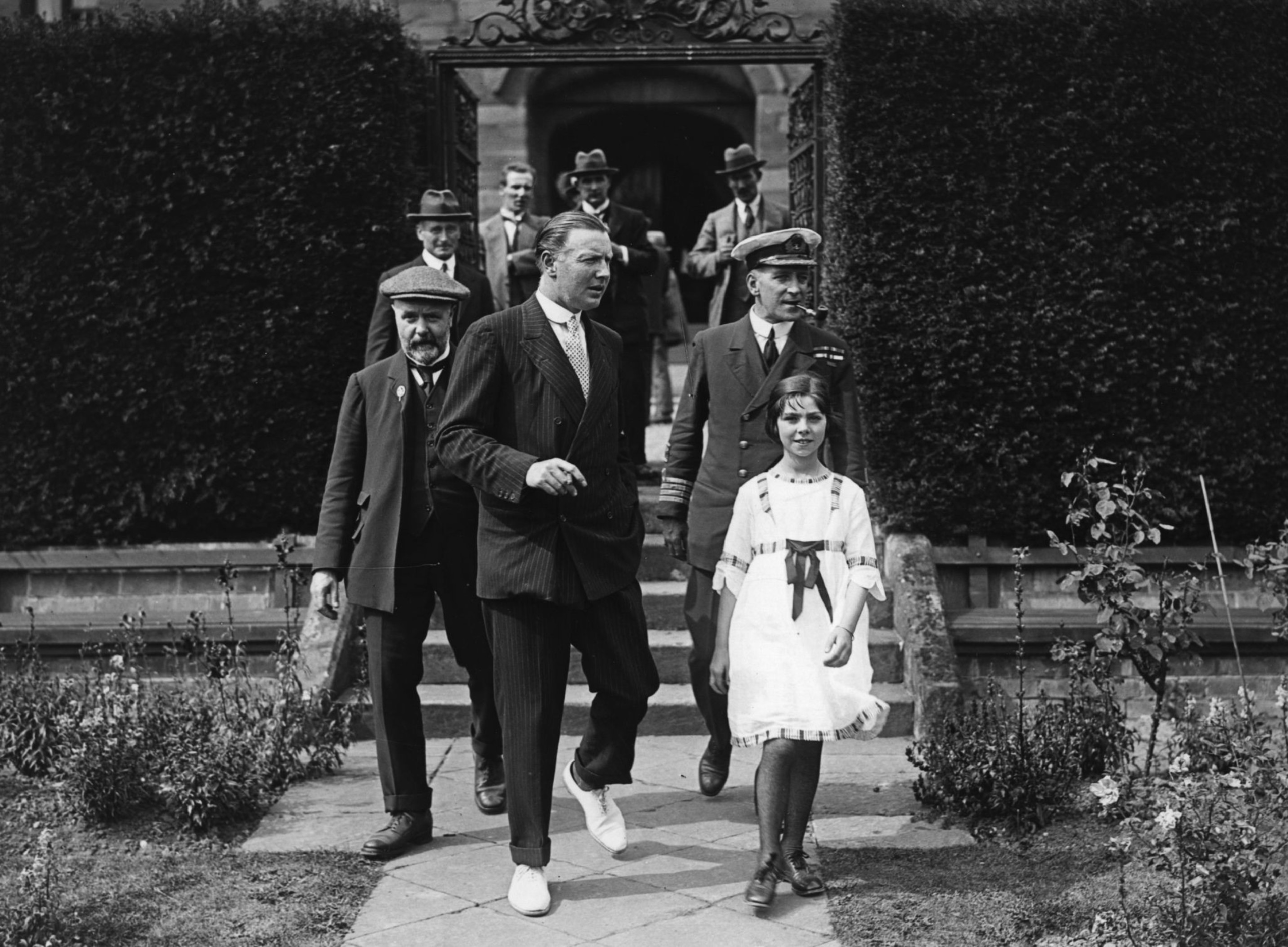 A group of men in suits and uniforms walk down garden steps behind a young girl in a white dress with a dark sash, framed by hedges and flowers.