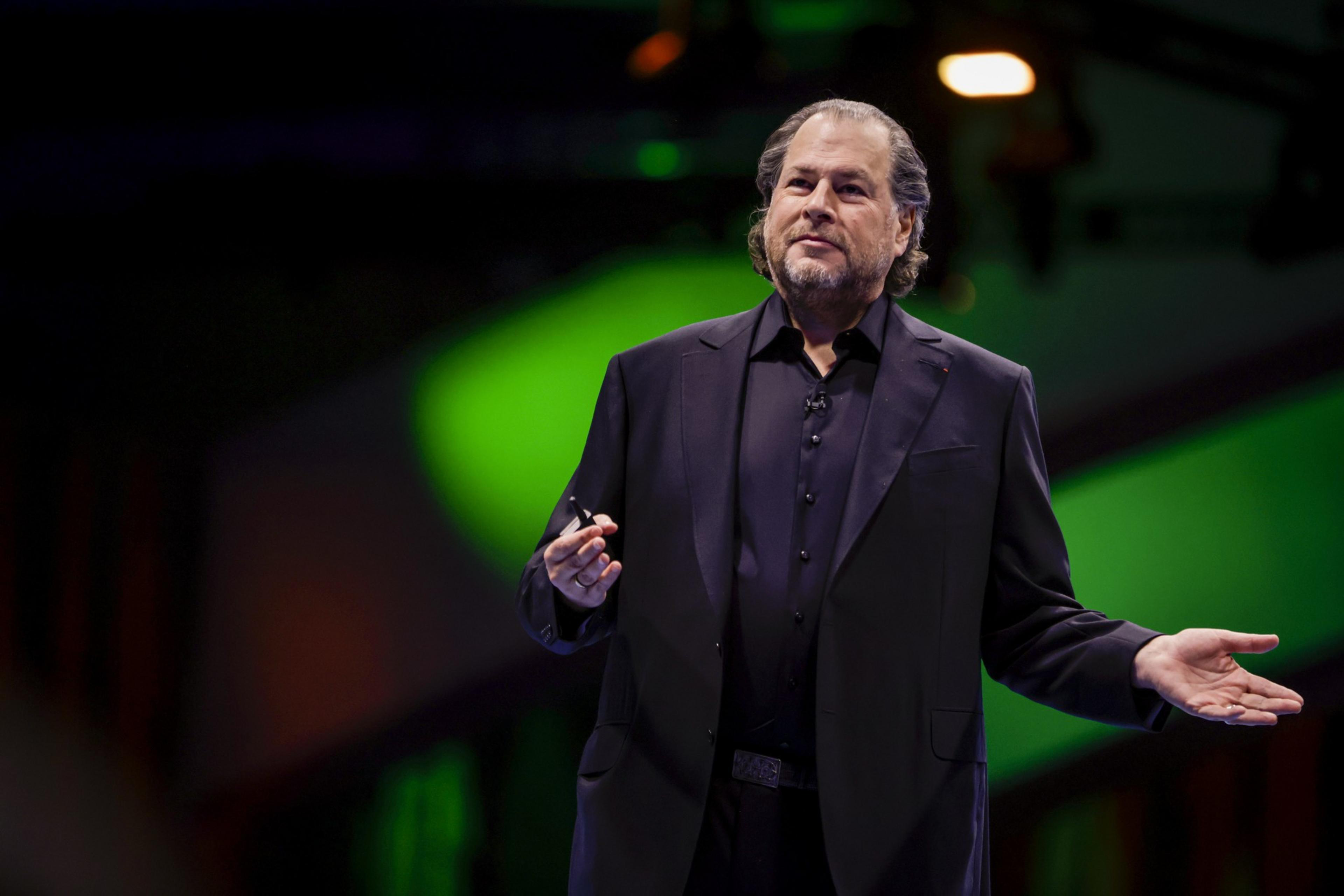 A man wearing a dark suit and black shirt gestures with both hands while speaking in front of a blurred green and dark background.