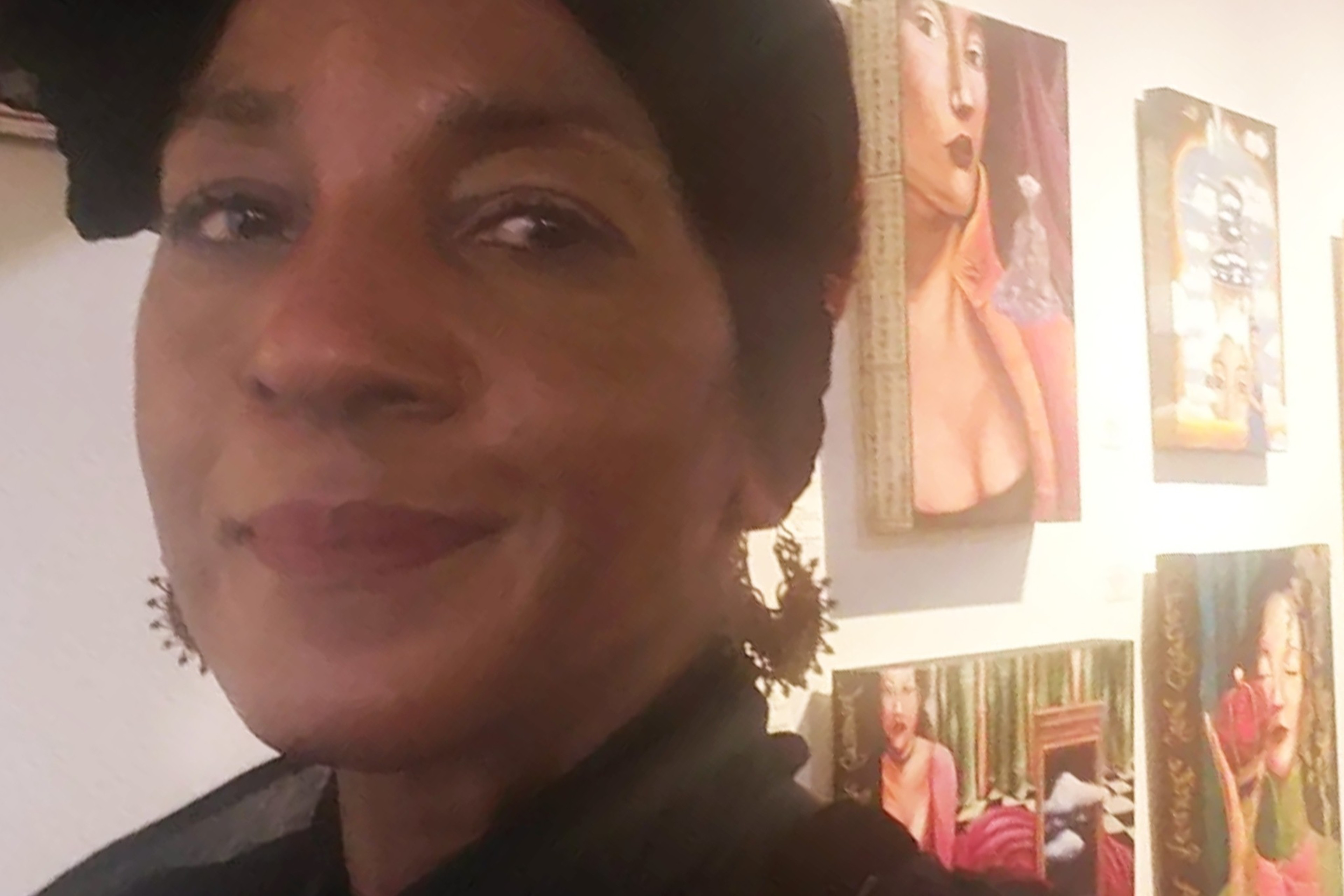 A woman with a headscarf and earrings is smiling slightly, standing in front of a wall displaying various colorful paintings, including portraits of women.