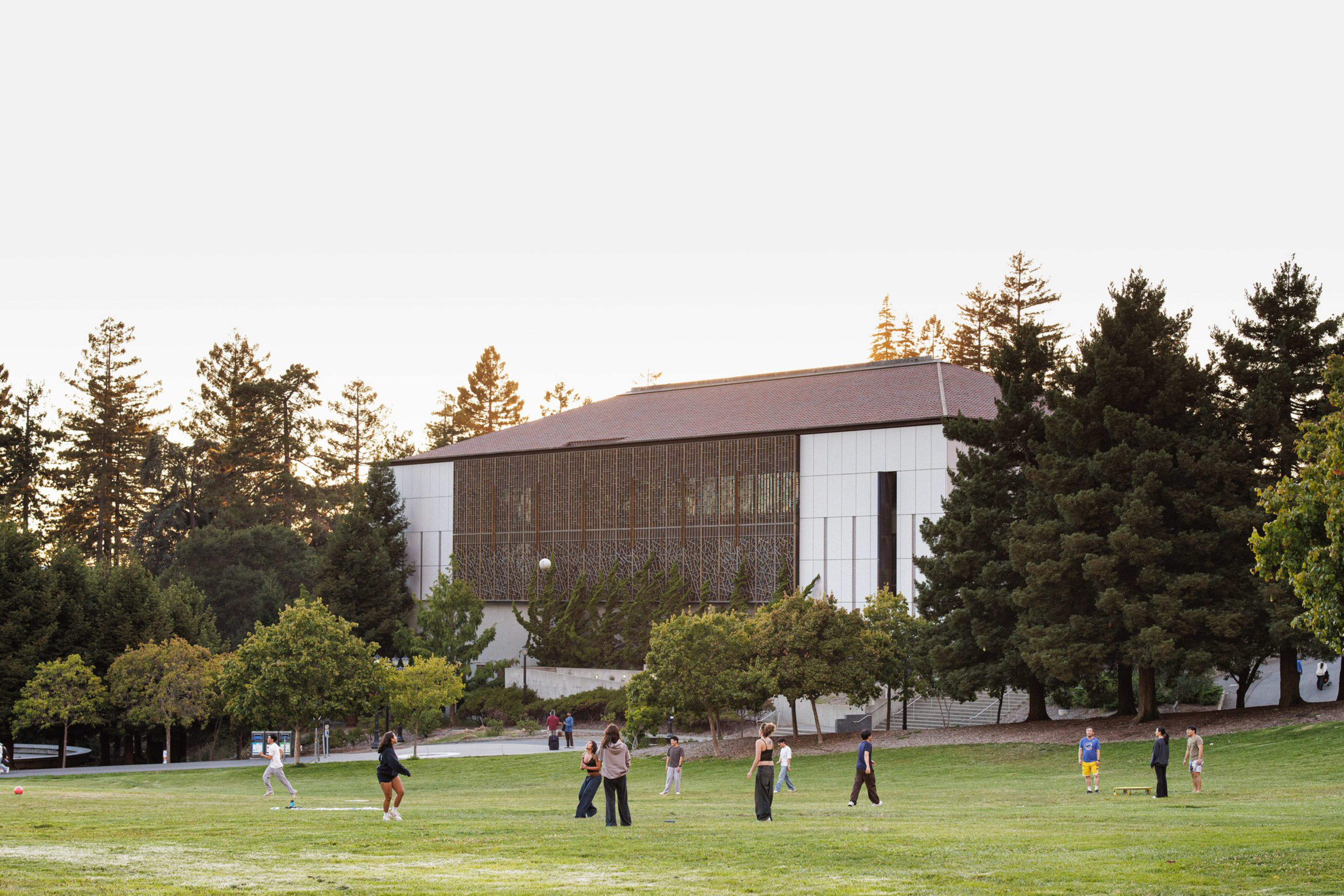 People are scattered across a grassy lawn playing and walking near a modern building surrounded by tall trees under a clear sky.