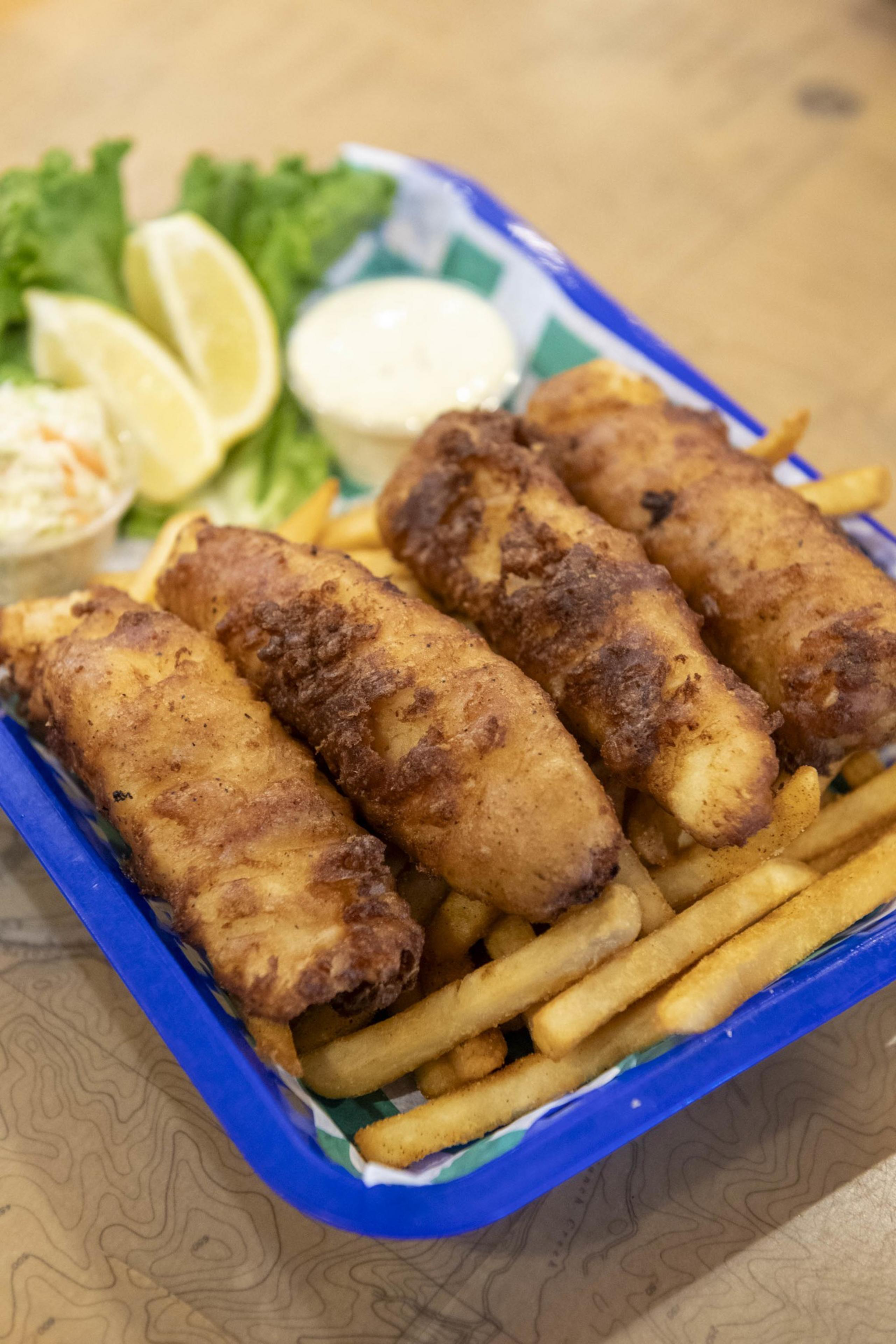 The image shows a basket of golden-brown fried fish on a bed of fries, with lemon wedges, coleslaw, and tartar sauce on the side.