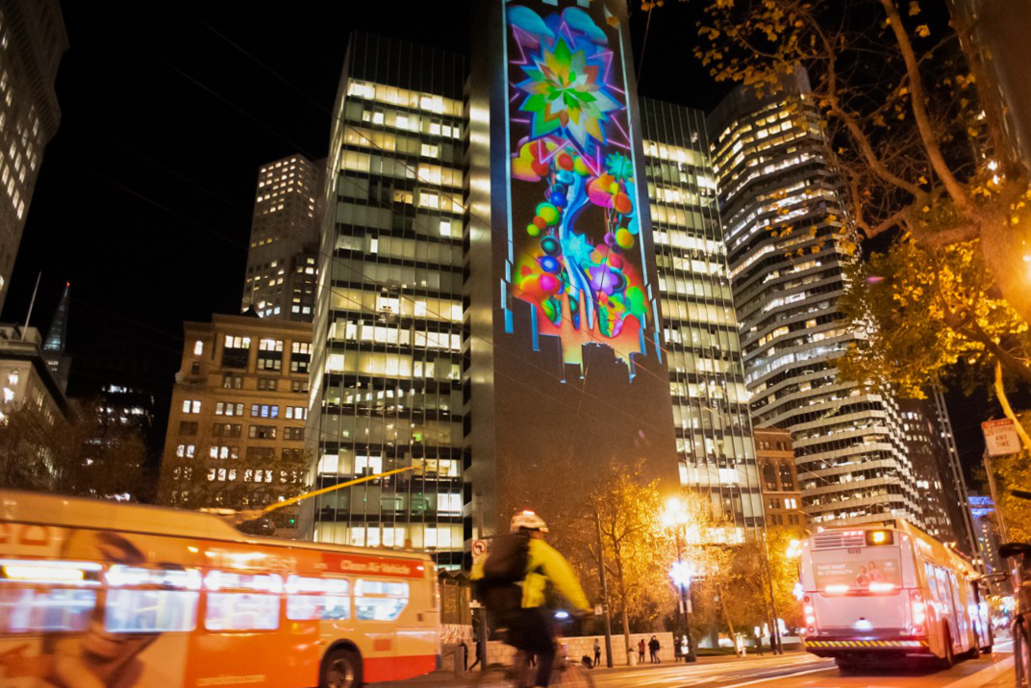 A colorful floral and geometric light projection adorns a tall building in a busy city street at night with buses and a cyclist passing by.