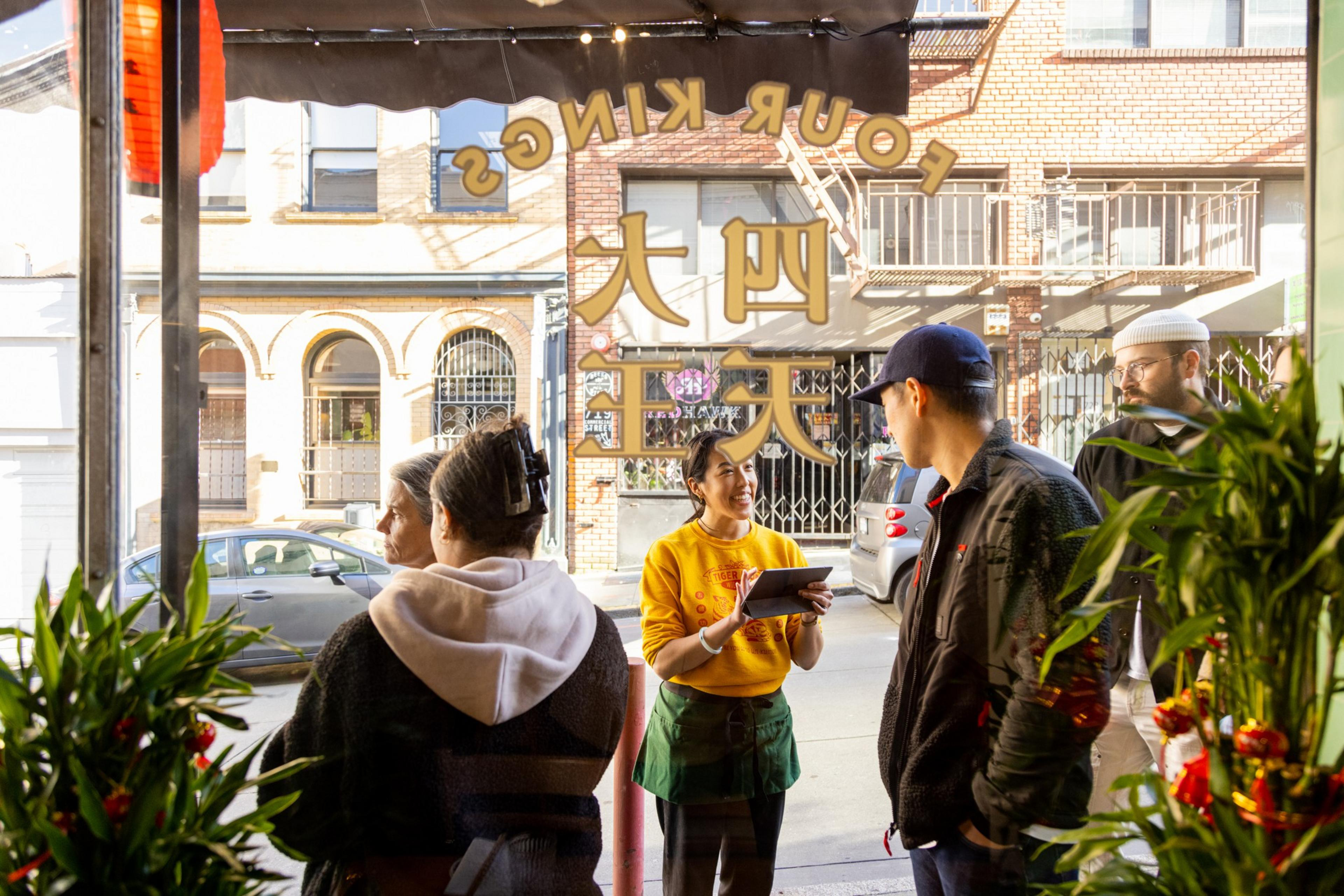 A waitress is taking an order from guests at a sunny outdoor restaurant entrance.