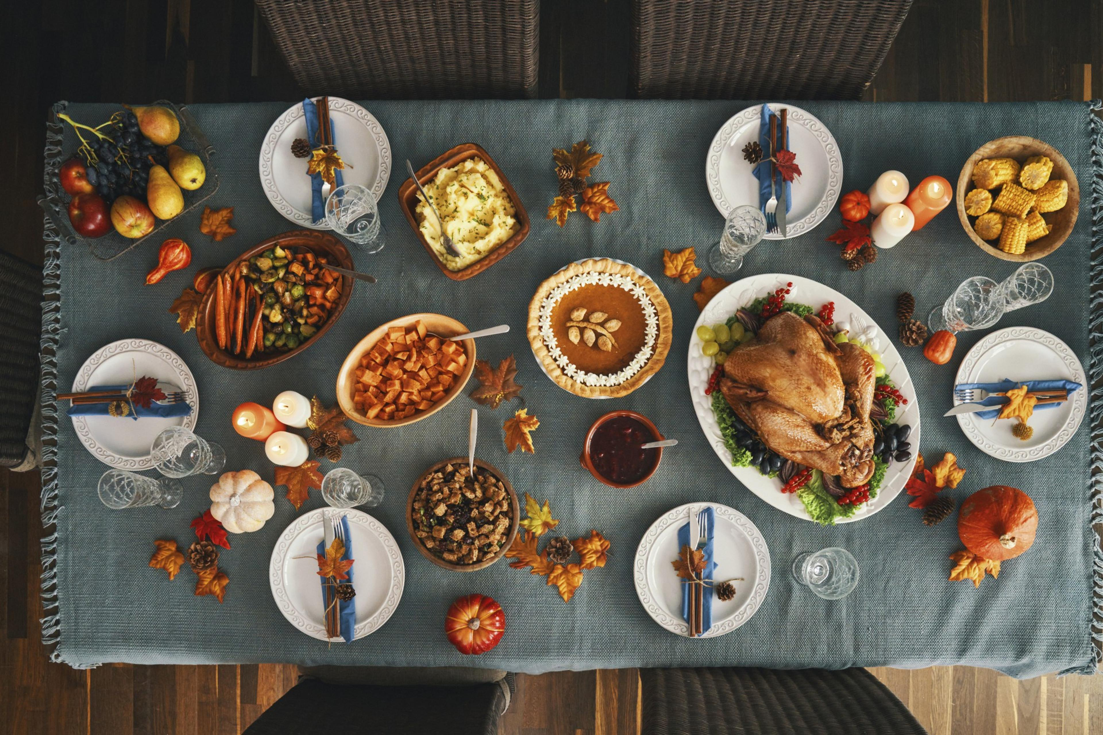 A festive dinner table is set with a roast turkey, pumpkin pie, various side dishes, fruits, candles, and autumn leaves on a blue tablecloth.