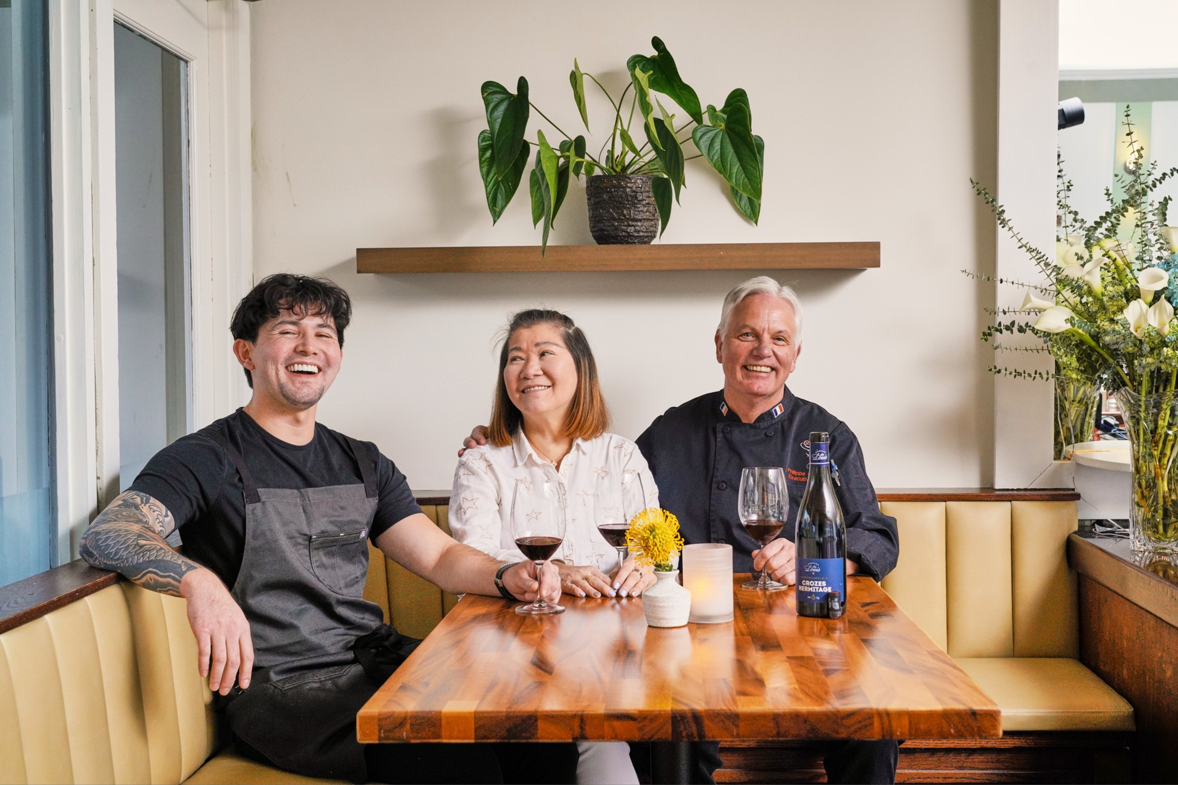 Three people sit at a wooden table with wine glasses, smiling. A plant is on a shelf behind them, and there are flowers on the table. The setting is cozy and bright.