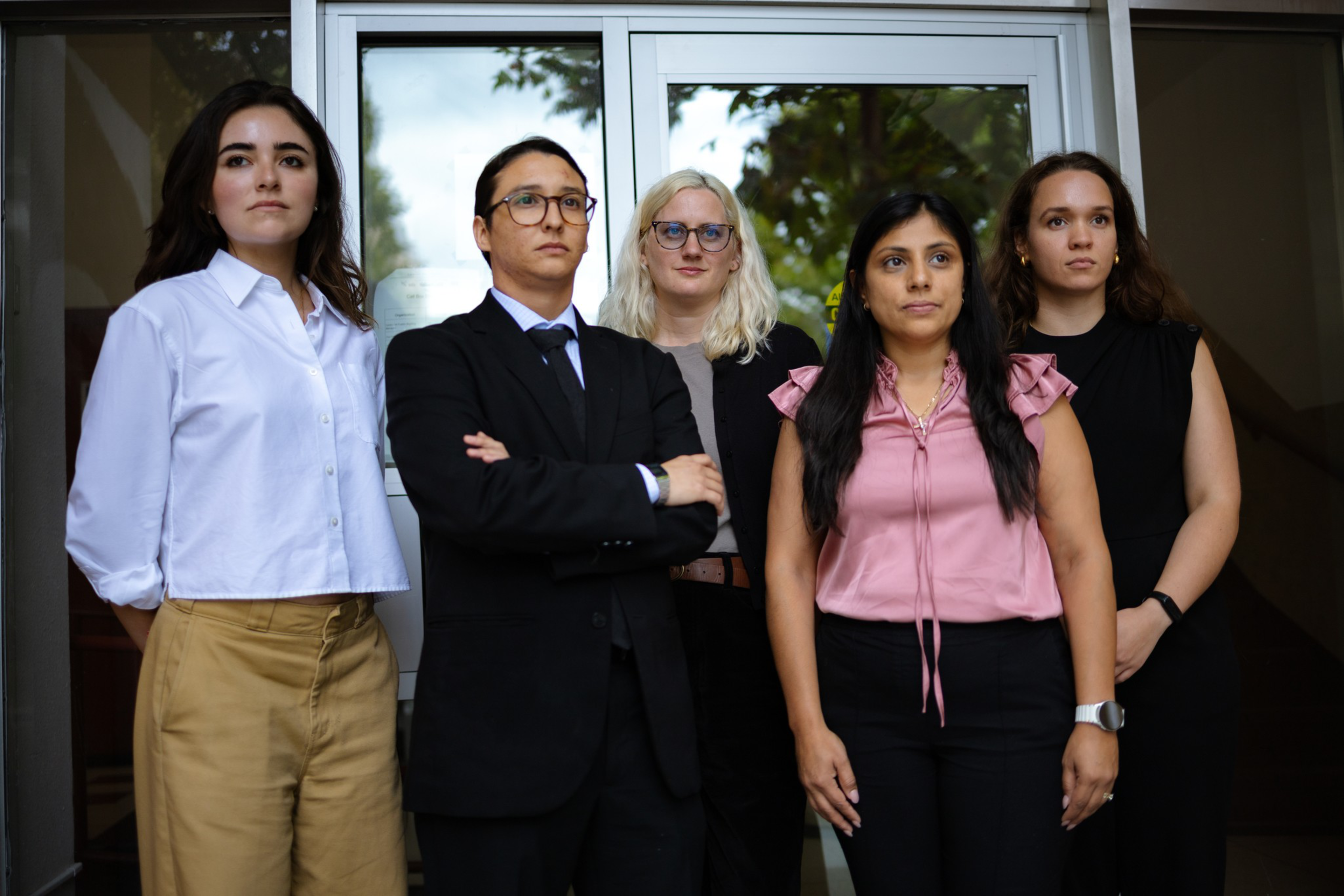 Five people stand in front of a glass door, looking serious; two wear glasses, one person is in a suit, and the others are dressed in business casual attire.